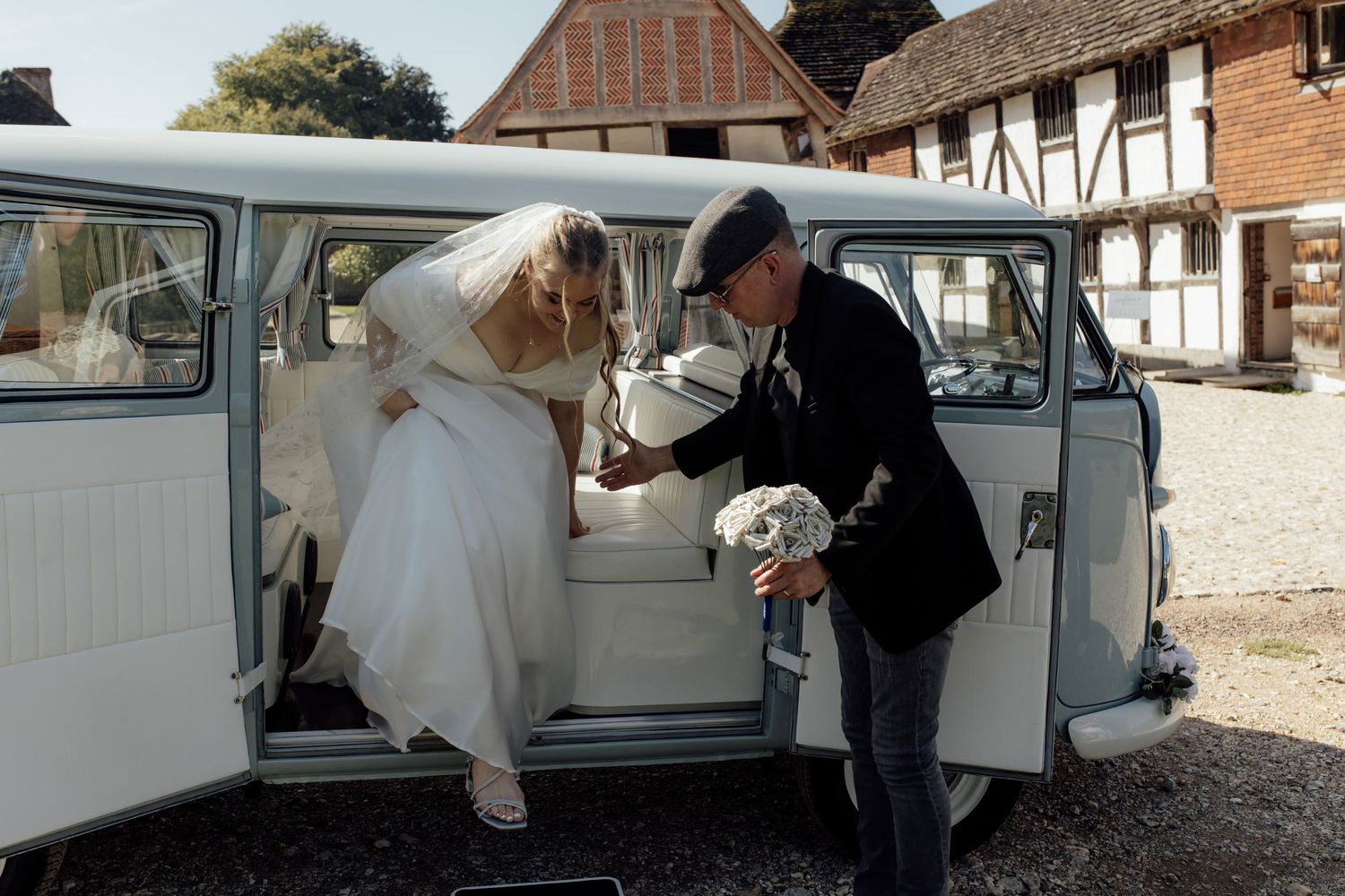 A bride in a white wedding dress gets assistance exiting a classic vintage car on her special day.