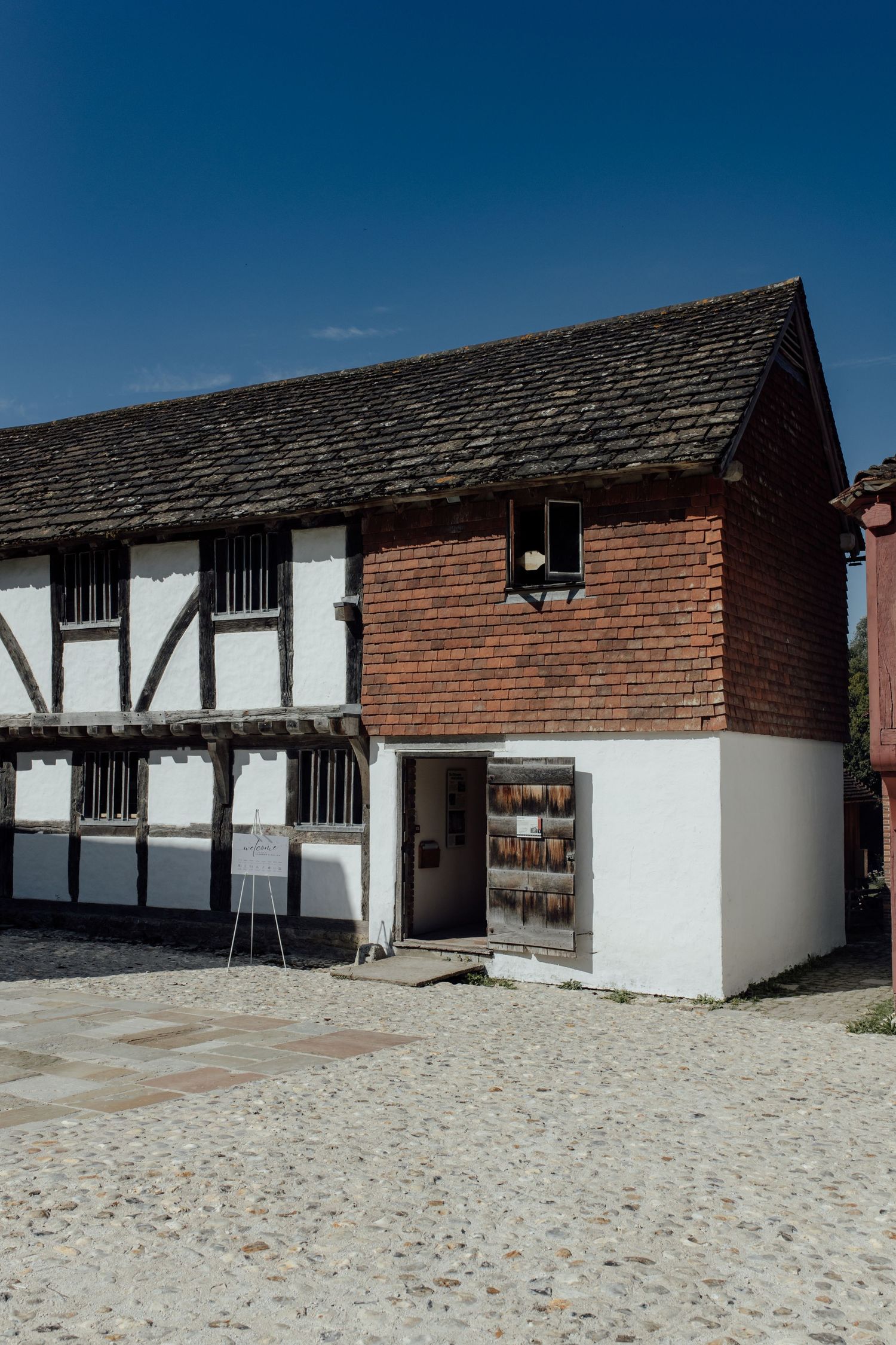 Historic Tudor-style building with white walls and red brick upper section features barred windows and sandy courtyard.