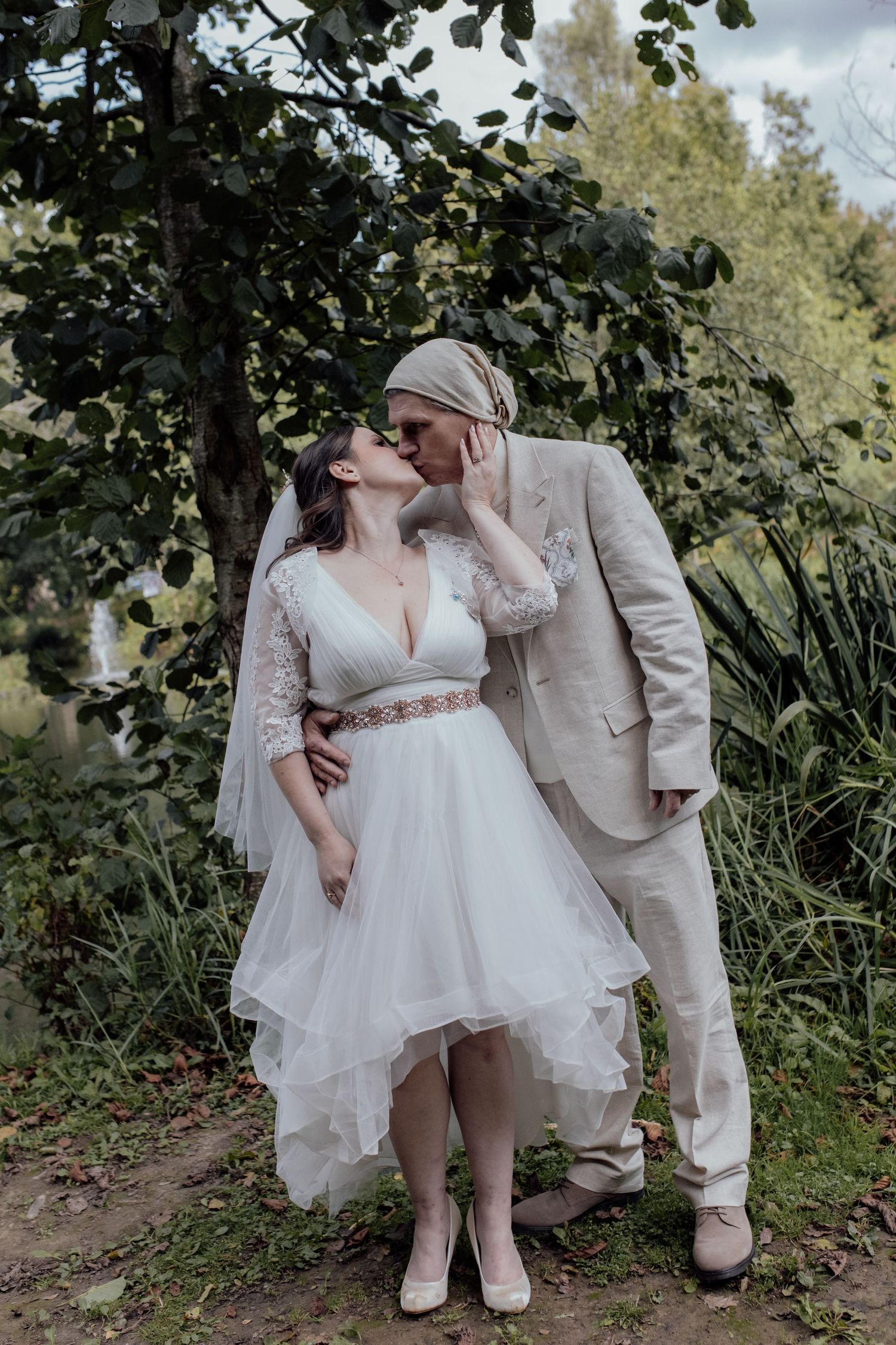 A romantic wedding couple poses together in a garden setting, with the bride wearing a flowing white dress and groom in tan suit.