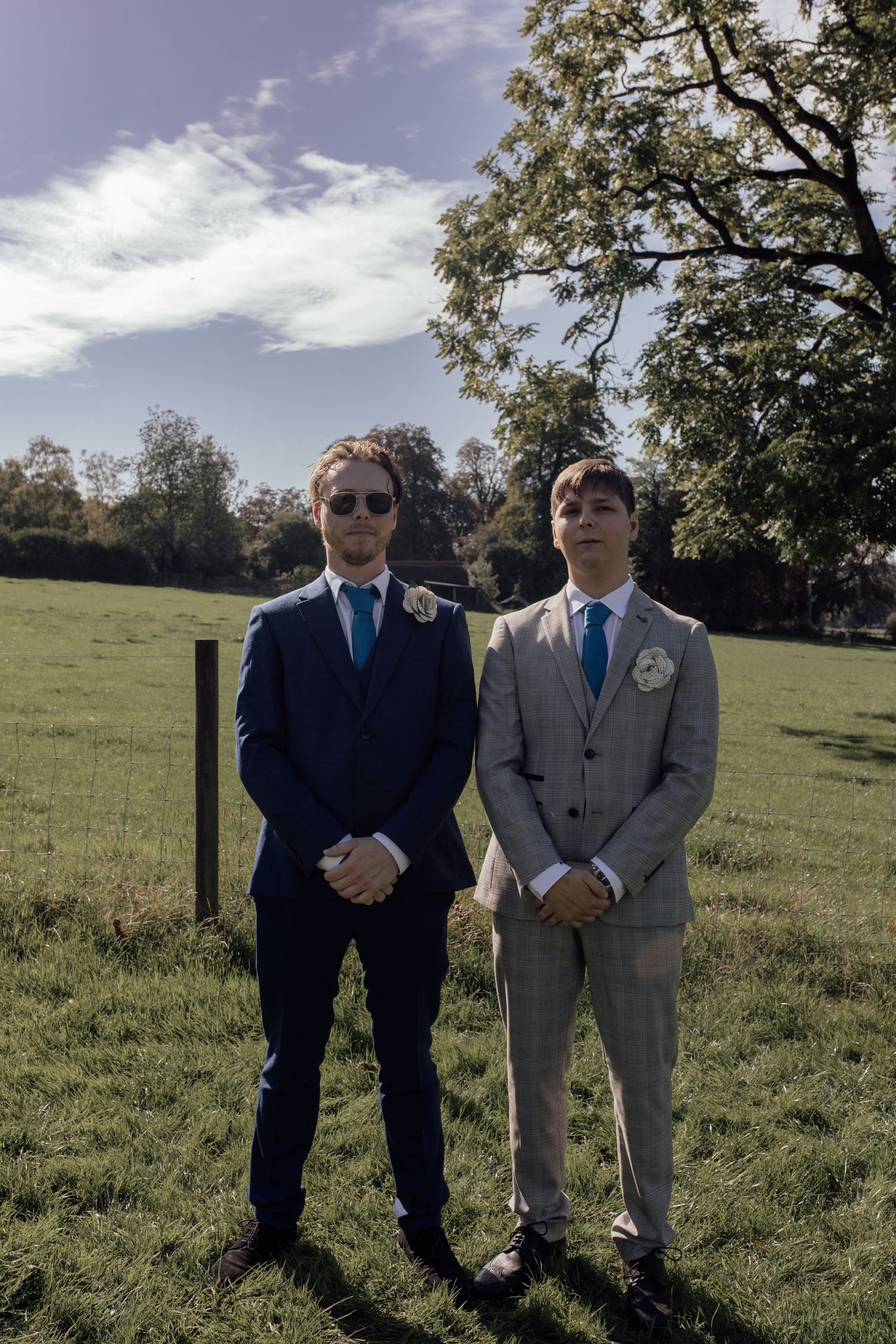 Two men in formal attire, one in navy and one in gray, stand together in a sunny grassy field.