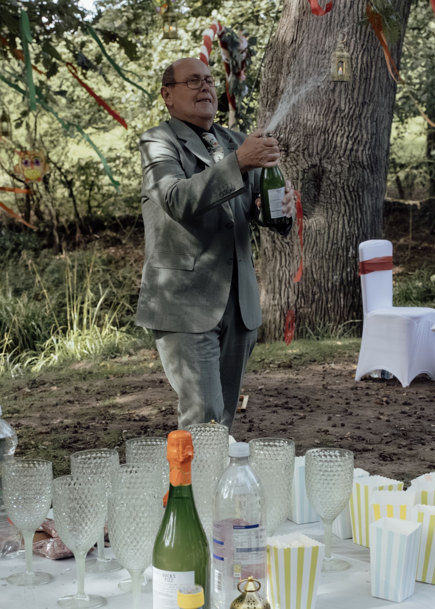 Someone in a gray suit pours champagne outdoors with wine glasses and party supplies set up on a table.