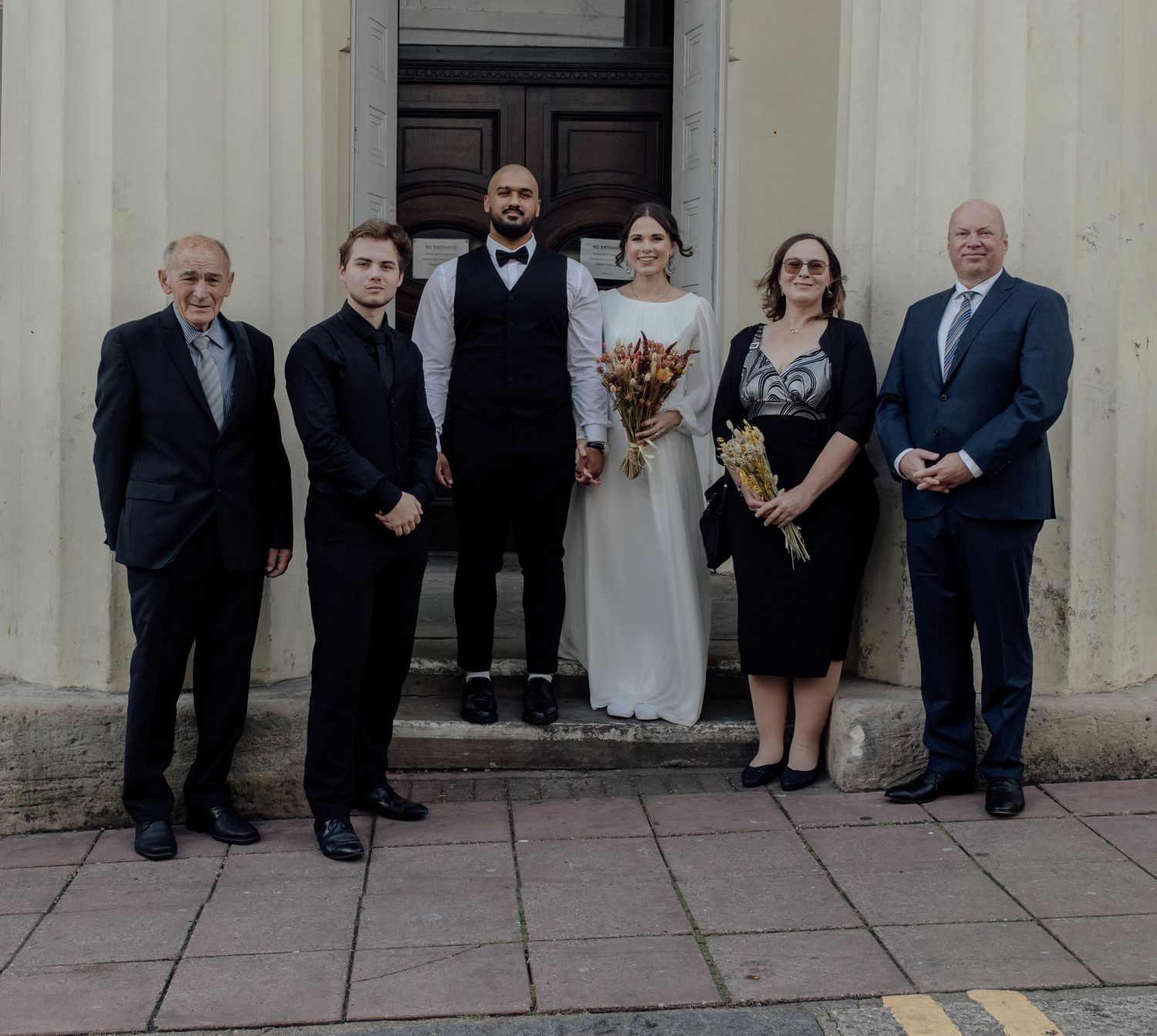 Wedding party stands in a row against a light colored building exterior.