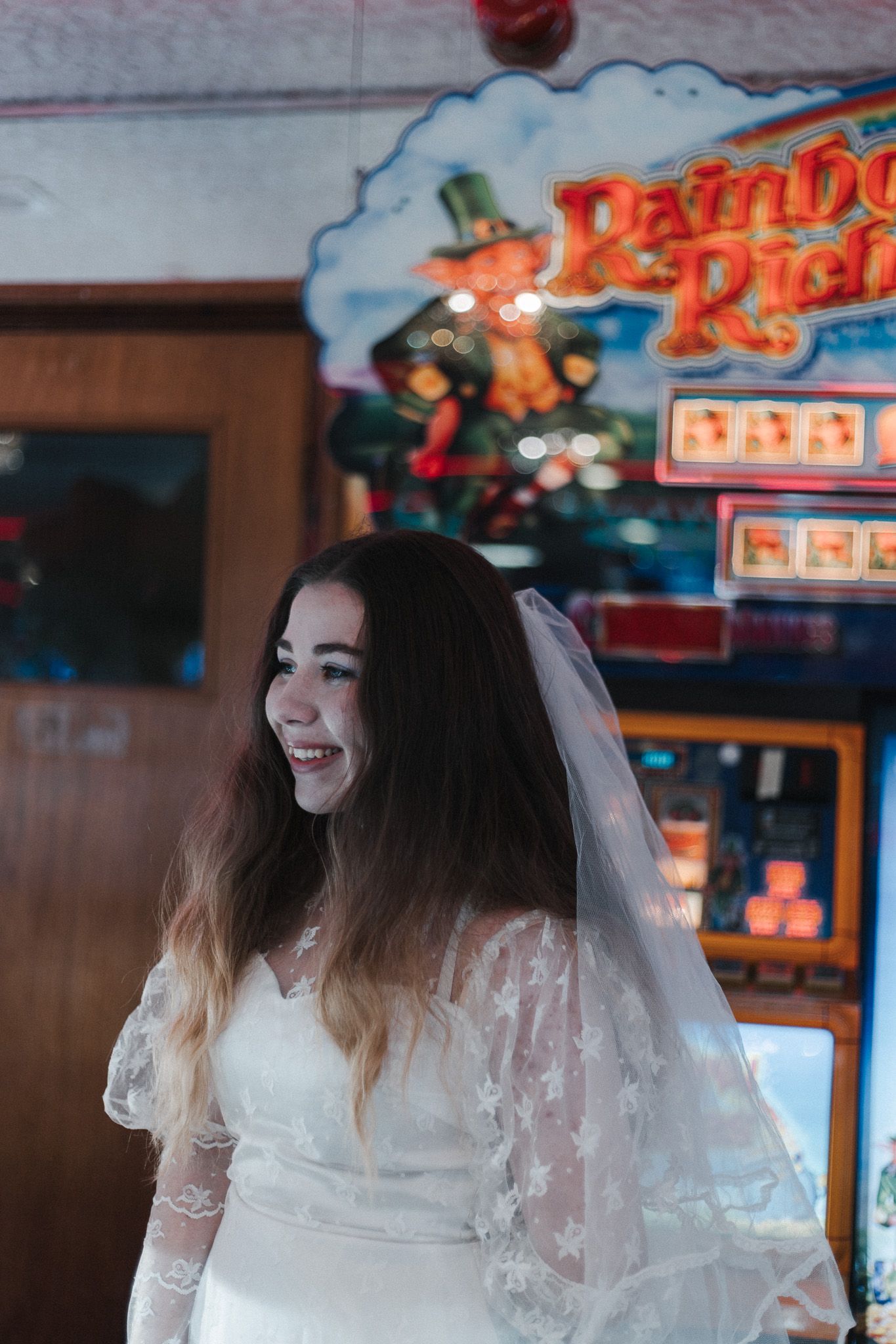 Bride in white lace dress laughs joyfully at an arcade with neon lights in the background.