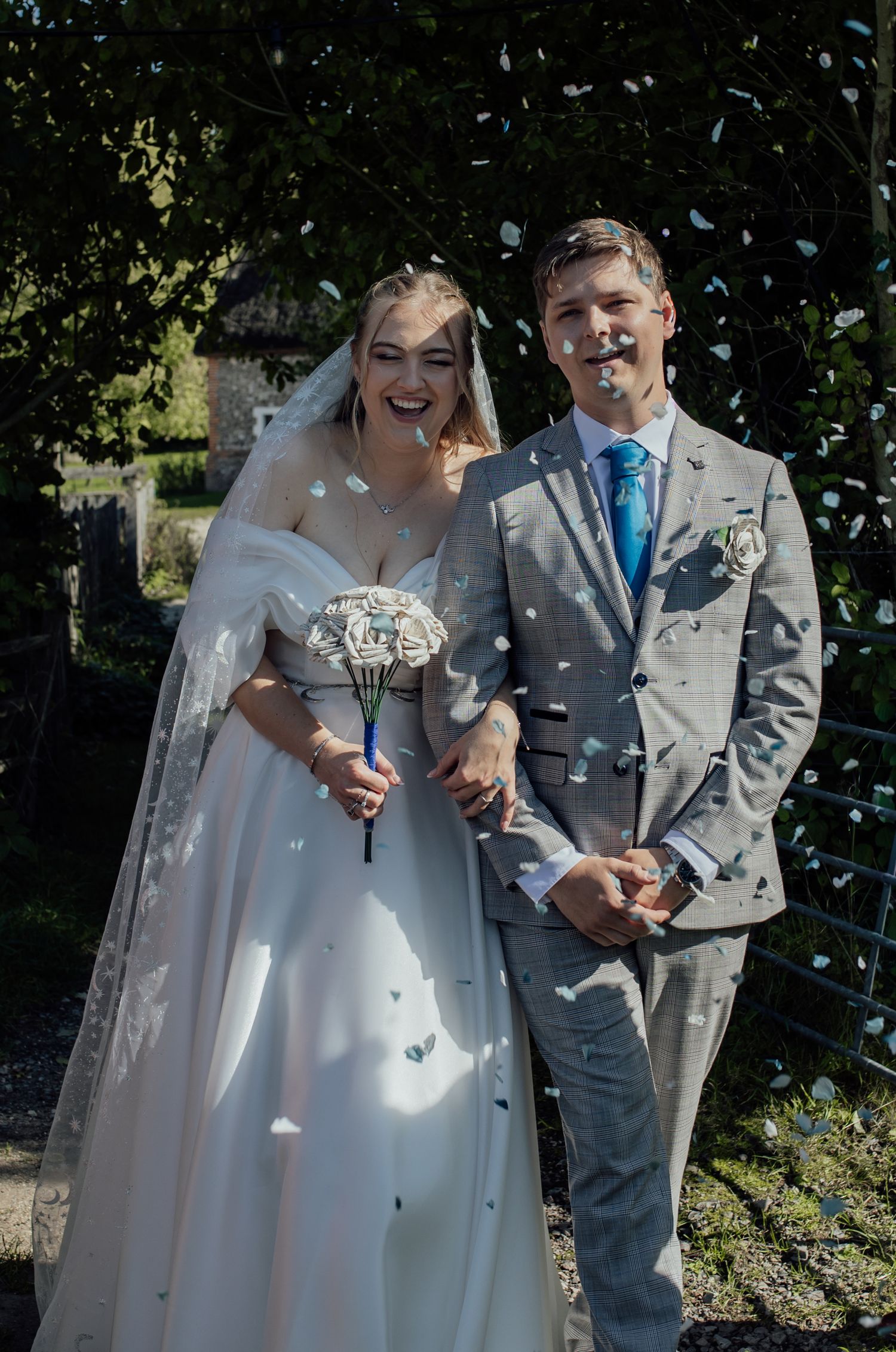 A bride in a white gown and groom in a gray suit stand together outdoors with confetti falling around them.