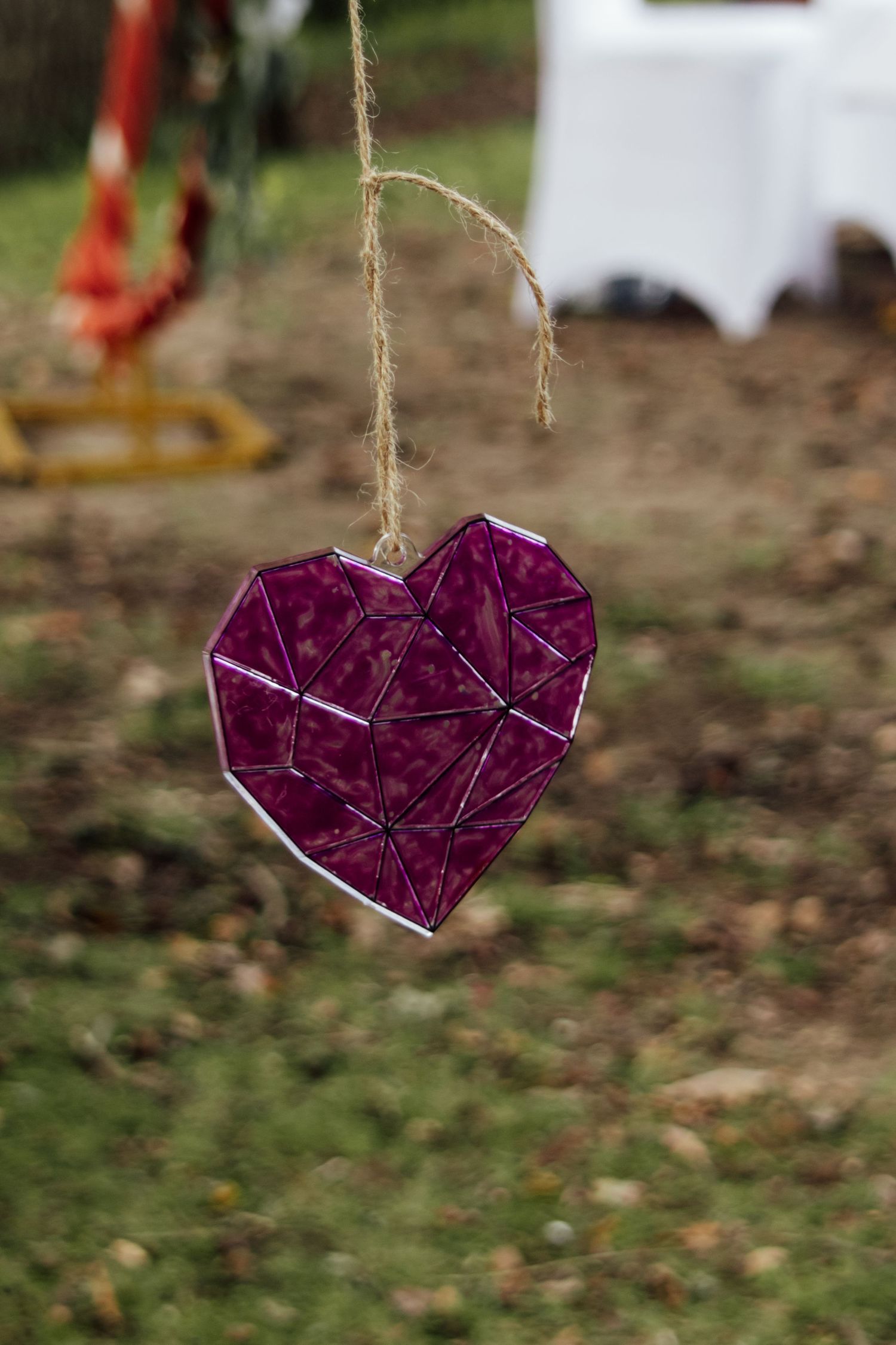 A purple heart-shaped ornament made of glass or crystal hangs from twine against a blurred outdoor background.
