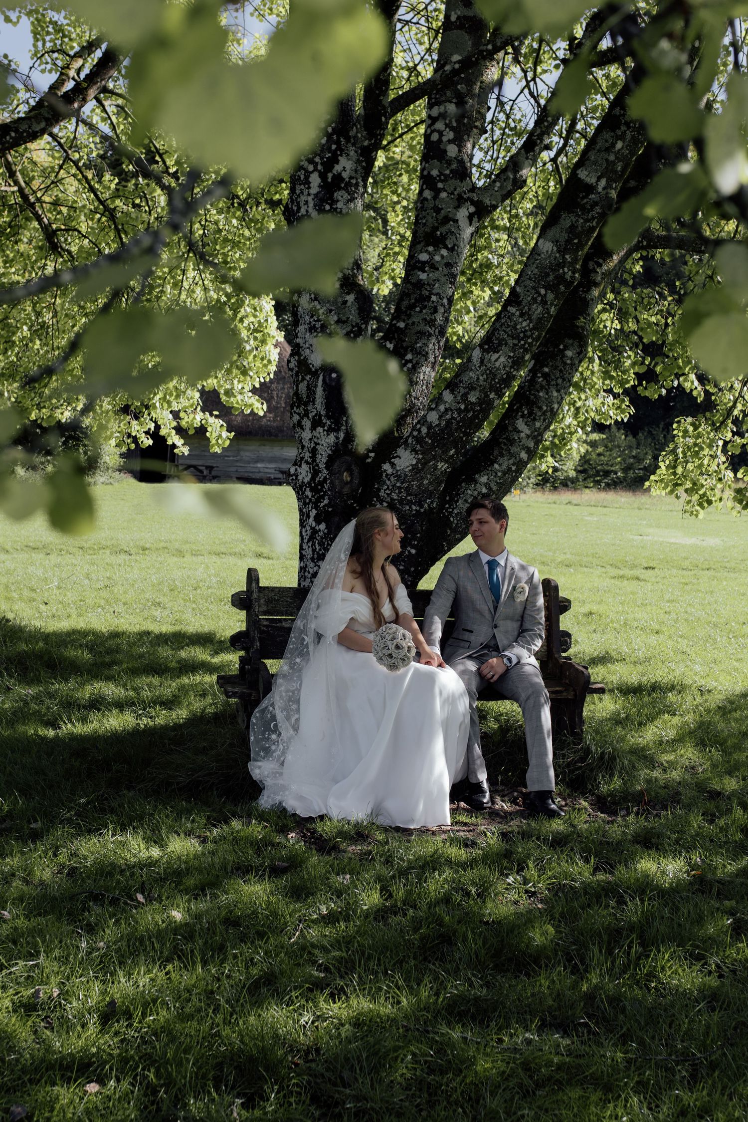 Couple sits together under a shady tree on a grassy hill during an outdoor wedding photoshoot.