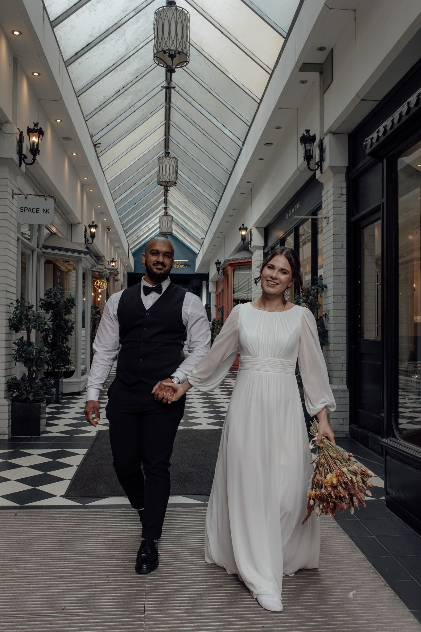 A couple in wedding attire walks through a bright indoor shopping arcade with a glass ceiling and black and white floors.