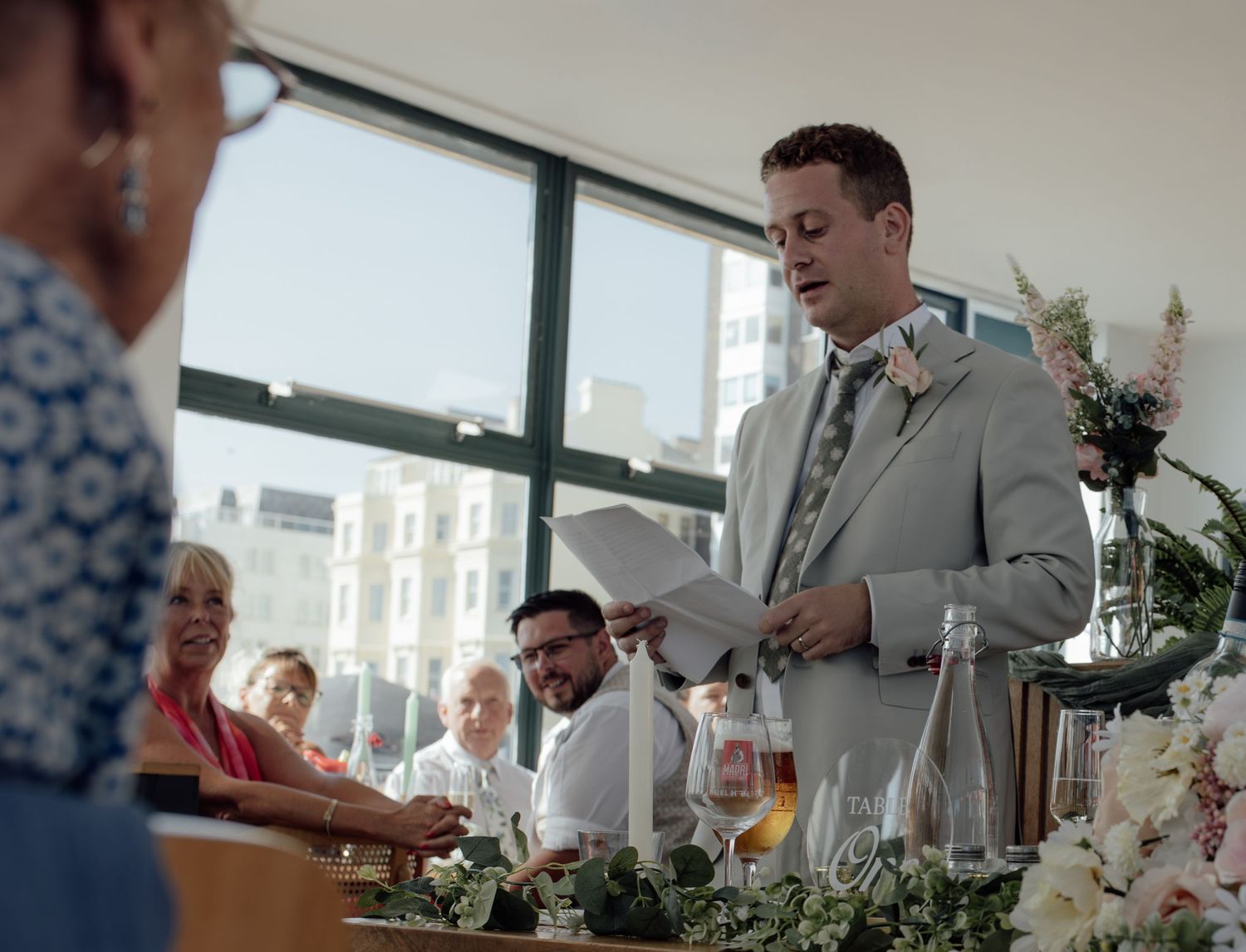 A speaker delivers a heartfelt speech at a wedding reception while guests listen attentively in a bright indoor venue.