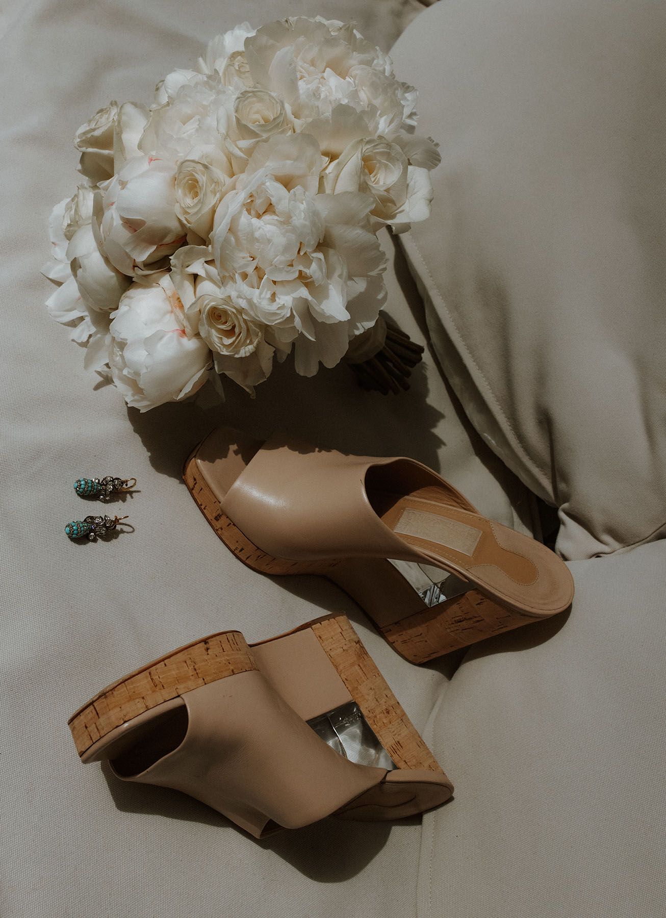 White rose wedding bouquet rests beside tan cork wedge sandals on light fabric.