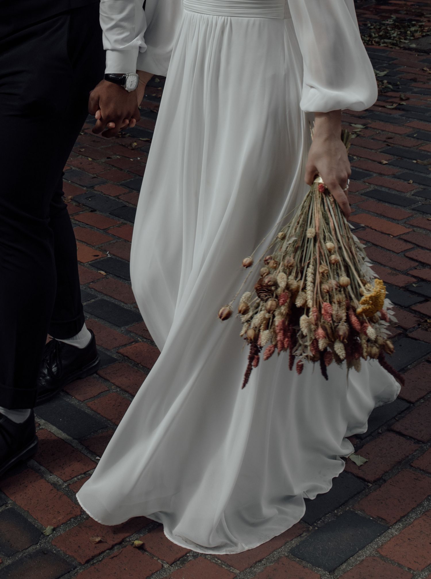 A wedding dress flows elegantly across brick pavement as the bride carries a dried floral bouquet in earth tones.