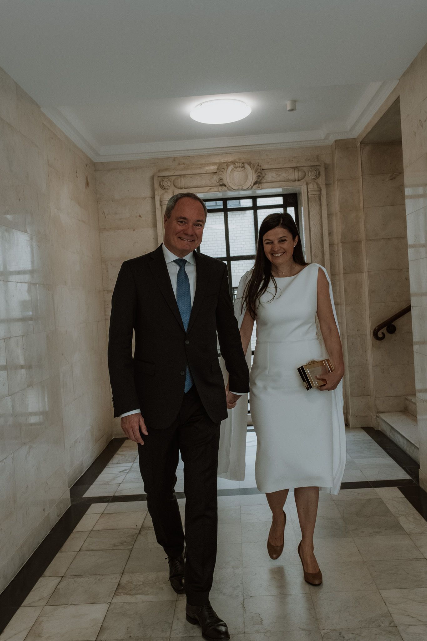 A couple walking down an elegant marble hallway in formal attire with natural light streaming through windows.