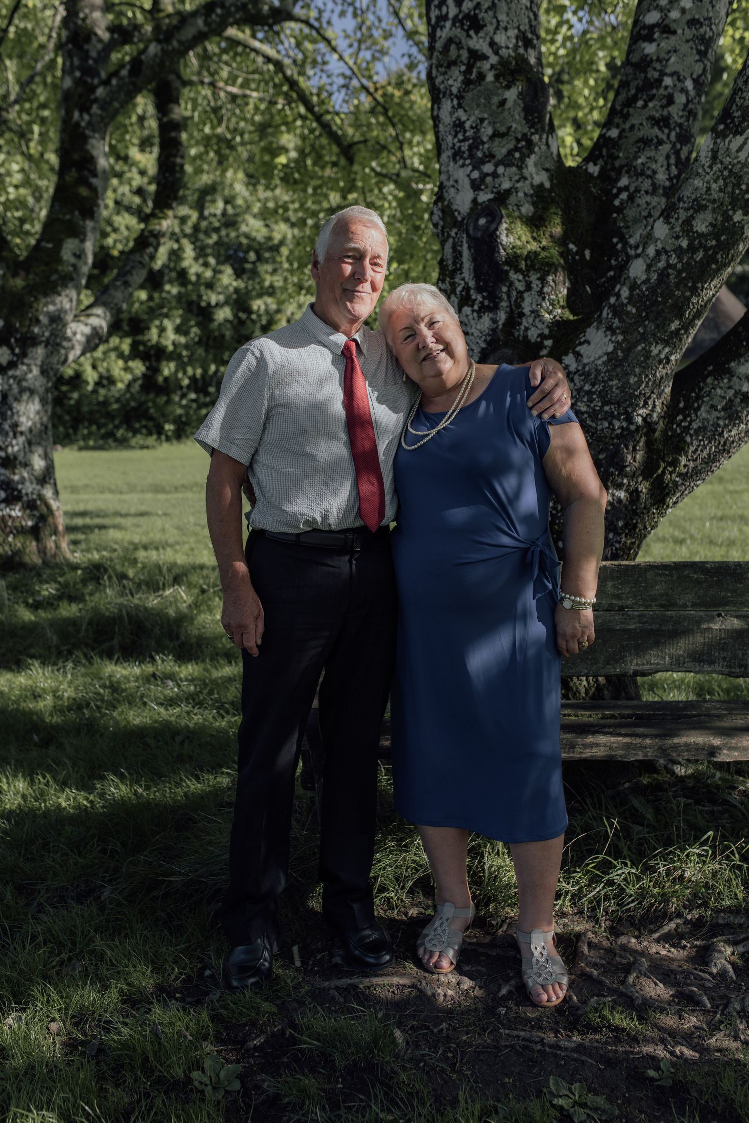 Two people stand together in a park setting among trees on a sunny day, casting shadows on the ground.