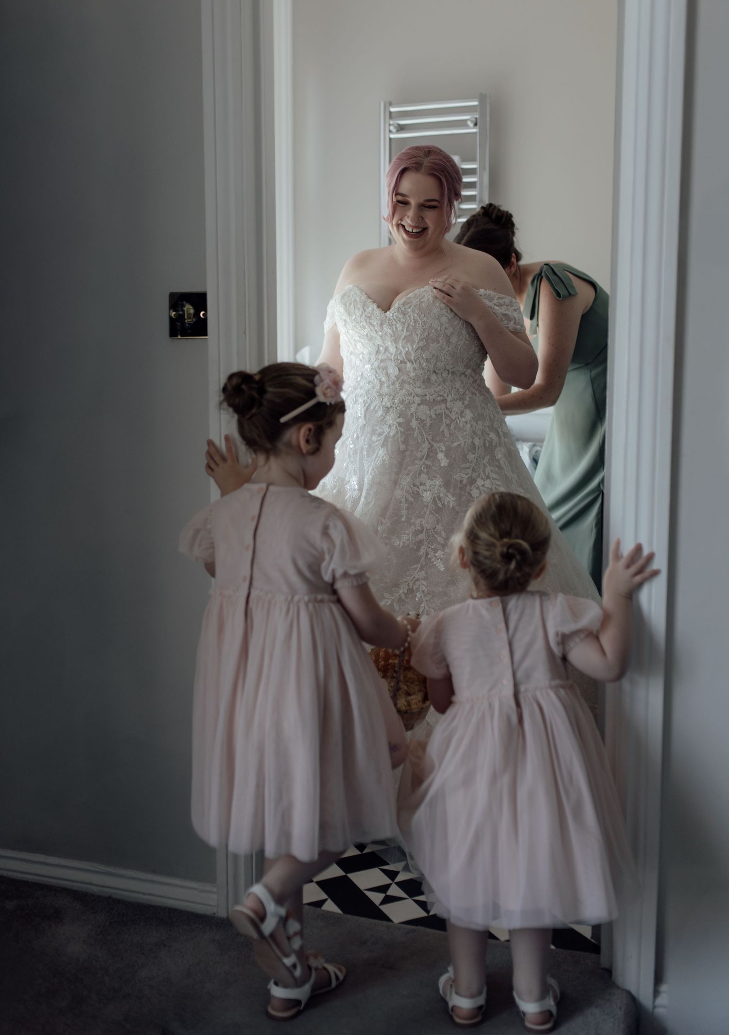 Two young children in pink dresses peek through a doorway while someone in a white gown stands nearby.