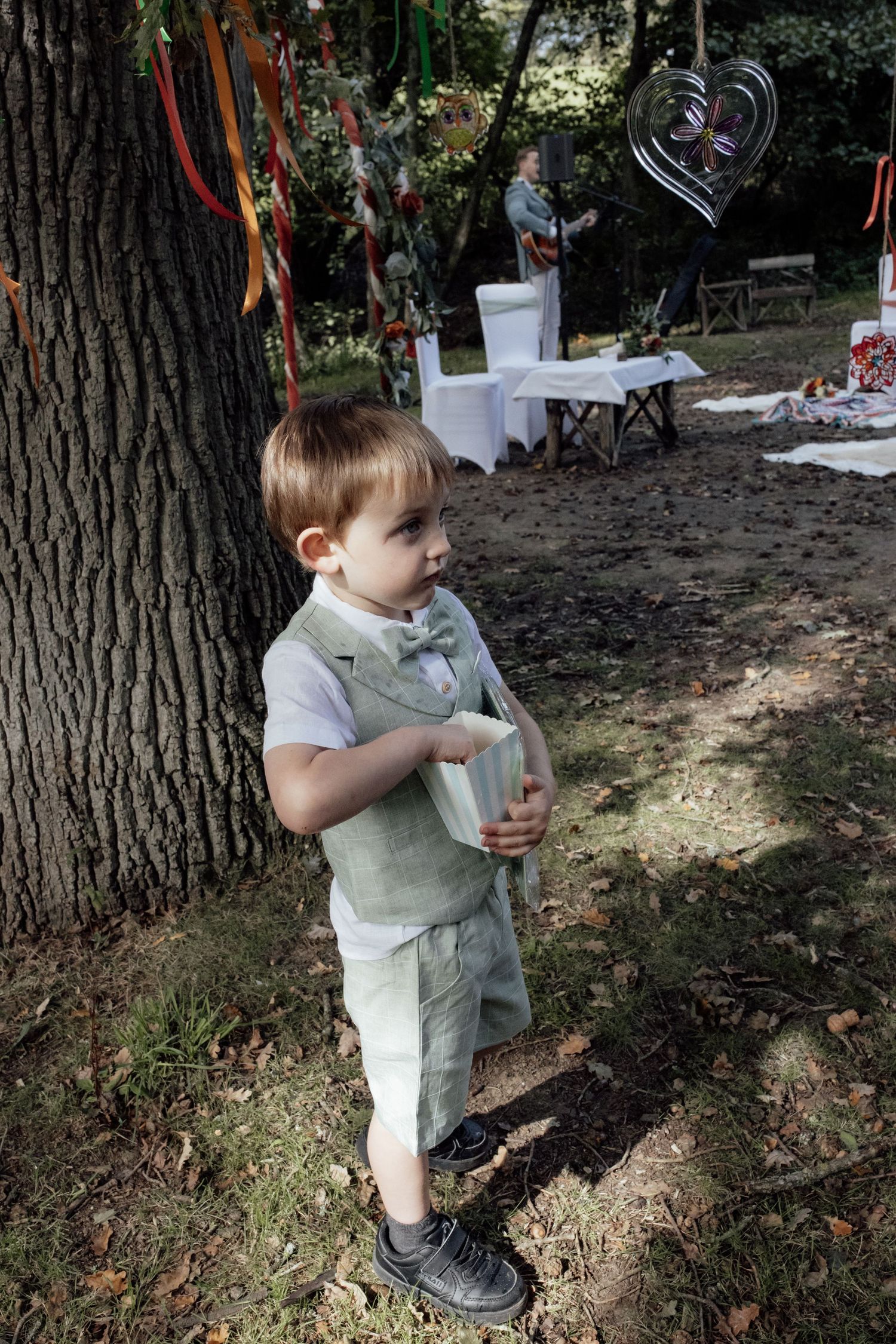 Young child in a light-colored shirt holding a drink cup while standing outdoors near trees in a park setting.