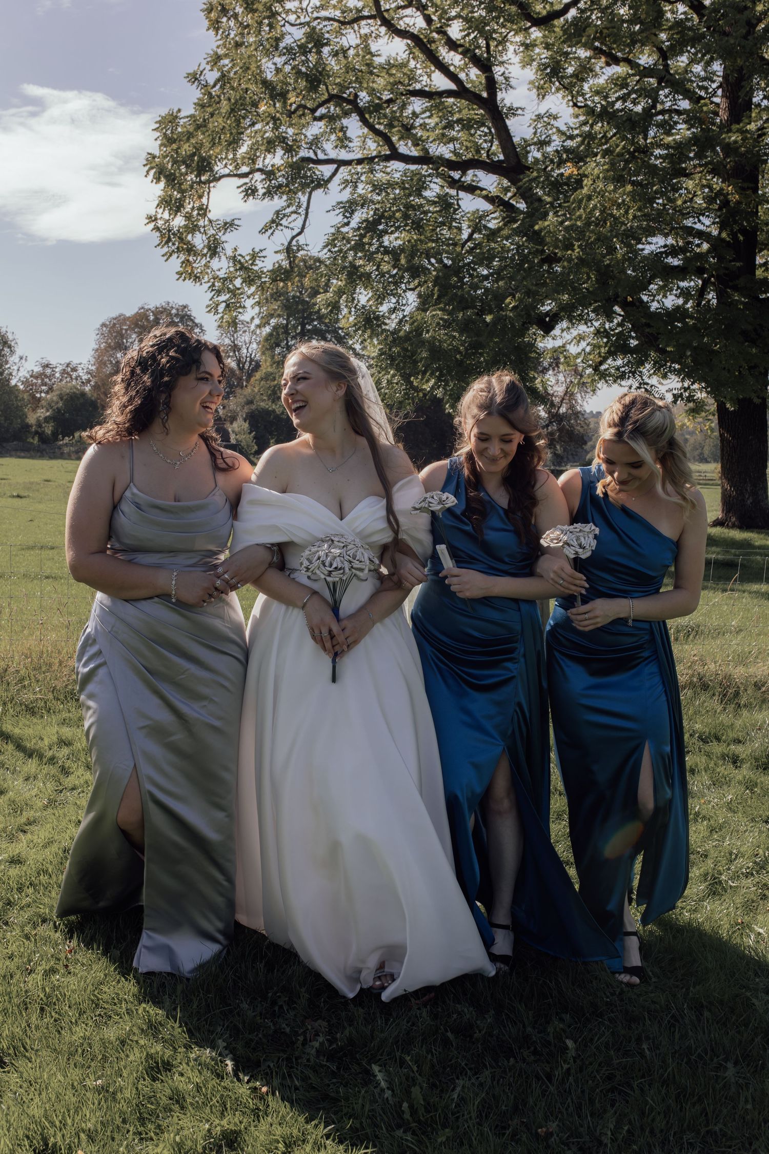 A bride in white gown walks with three bridesmaids wearing silver and blue dresses in a sunny outdoor setting.