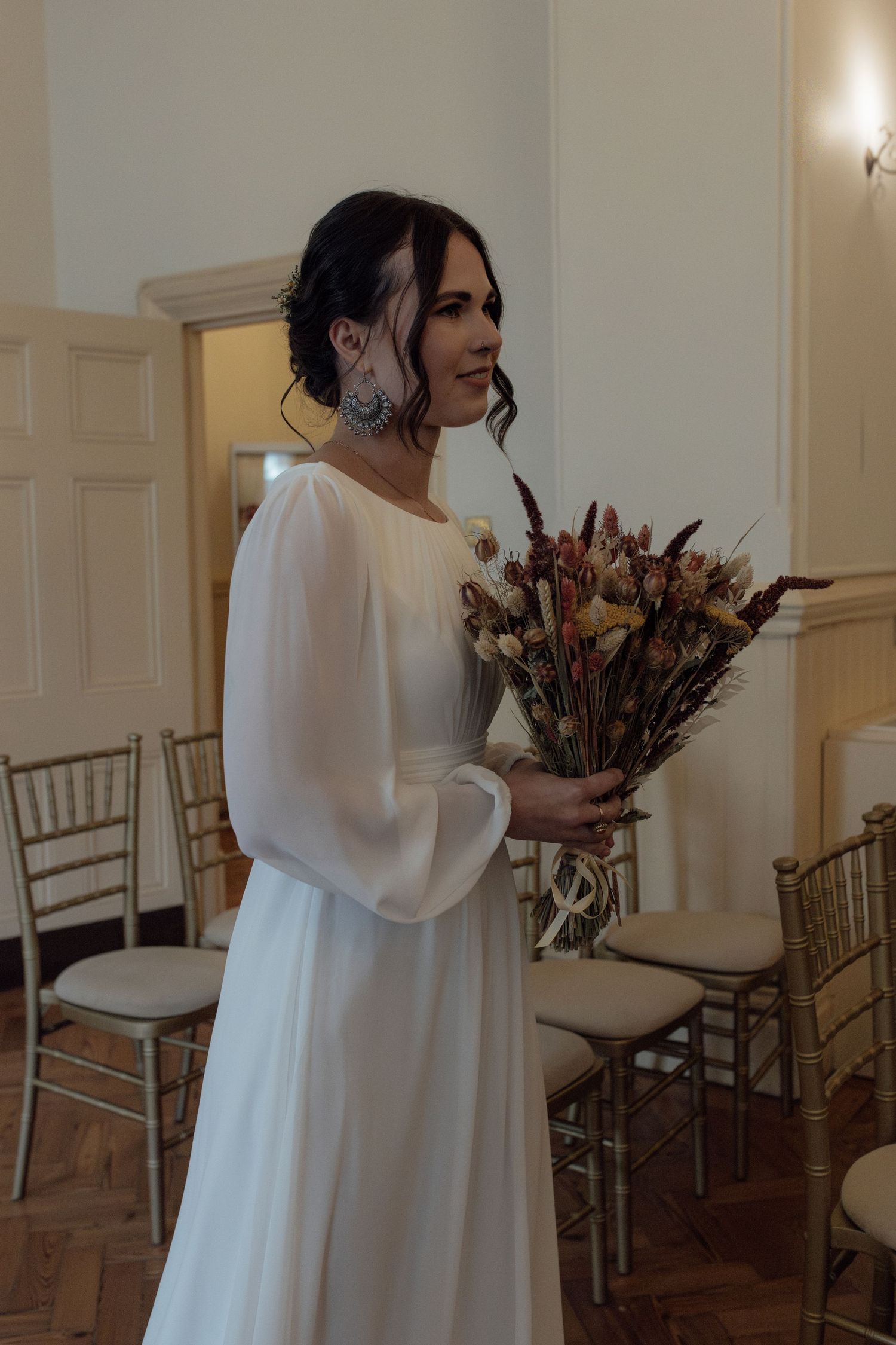 A figure in a flowing white dress holds a bouquet of dried autumn flowers in an elegant room with gold chairs.