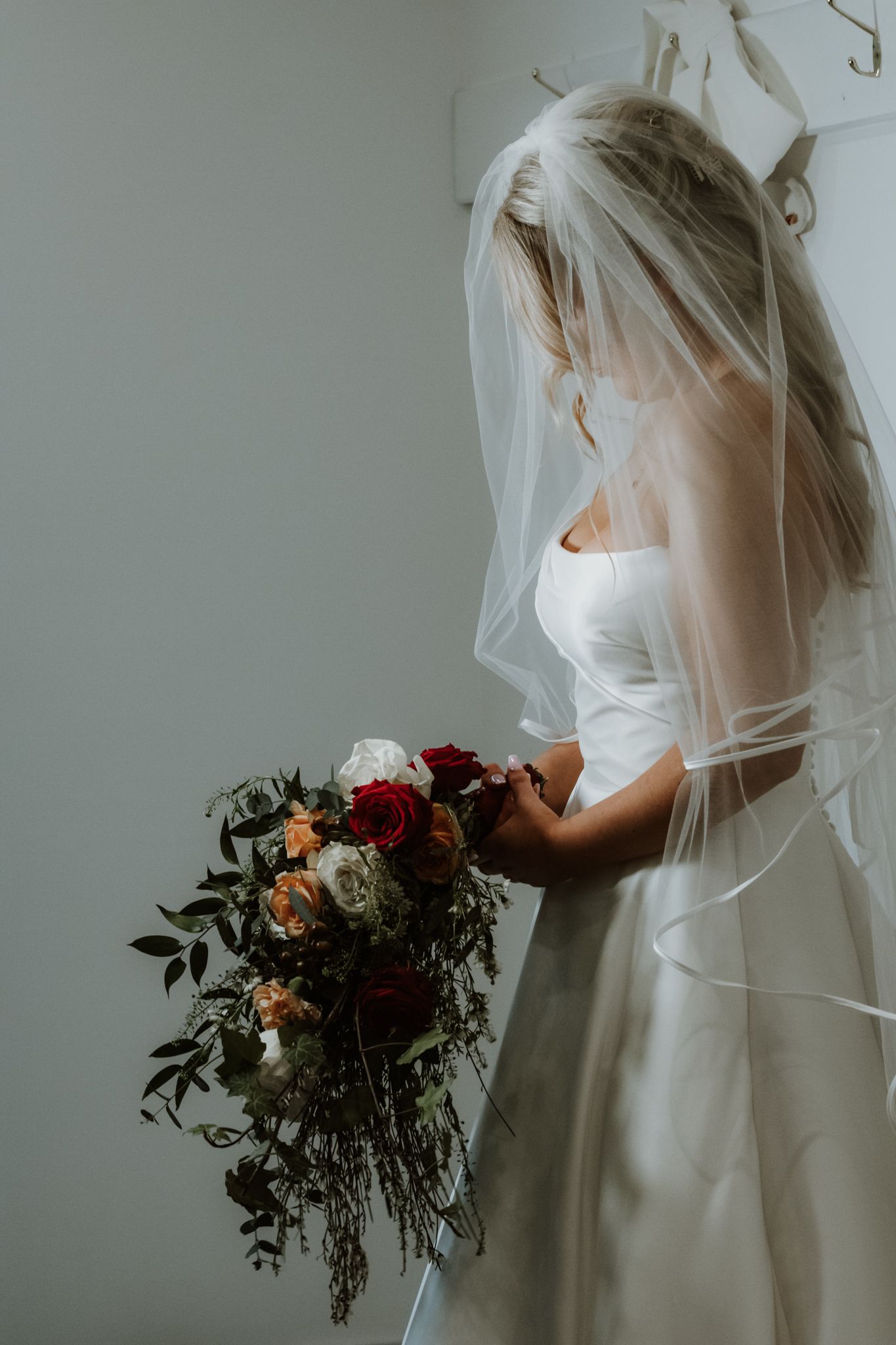 A bride in a white dress and veil holds a romantic bouquet of red and blush flowers against a light background.