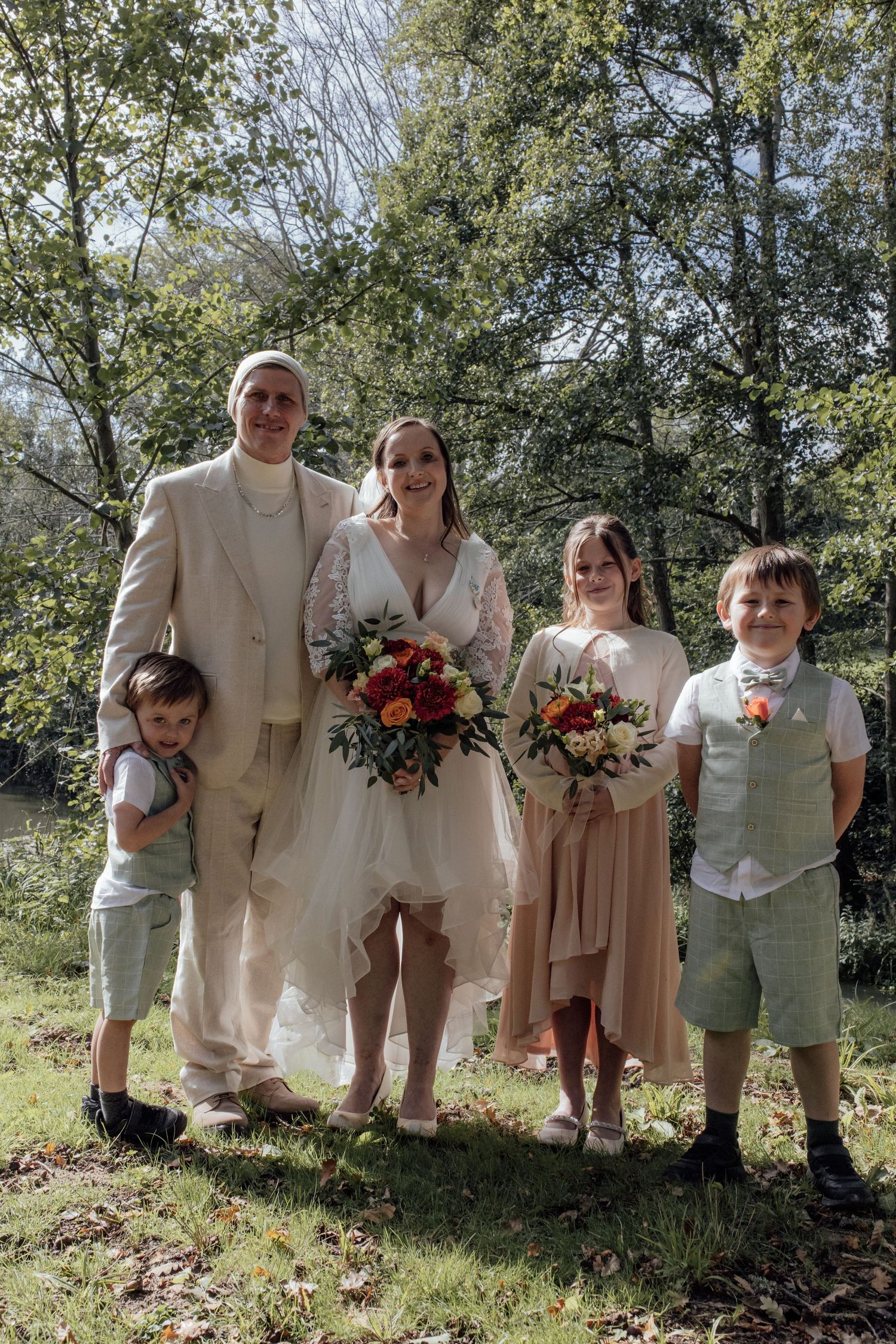 A group stands outdoors in light-colored attire at a wedding ceremony with autumn-colored floral bouquets.
