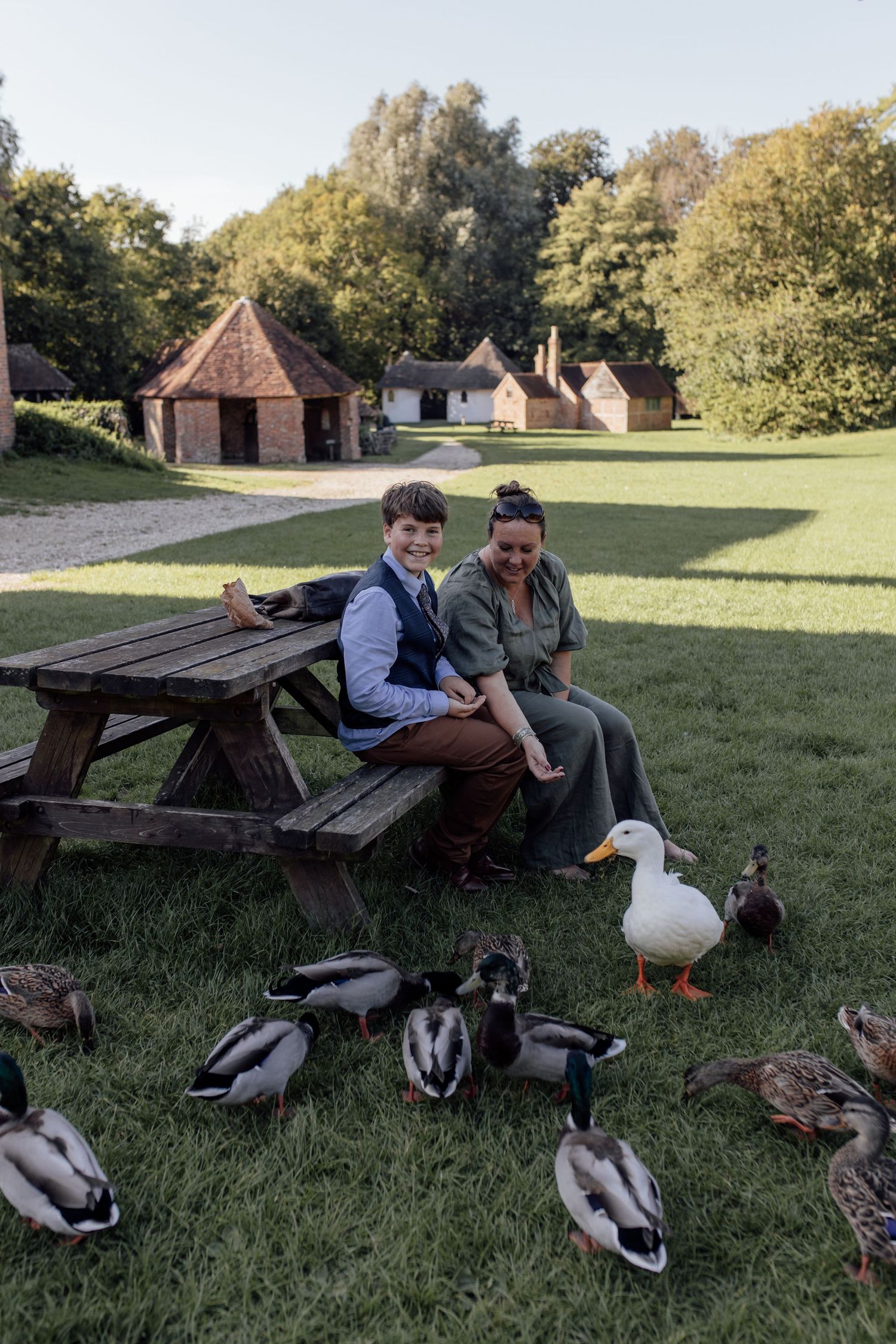 Historic colonial settlement with rustic buildings and picnic area surrounded by ducks and geese on grassy lawn.