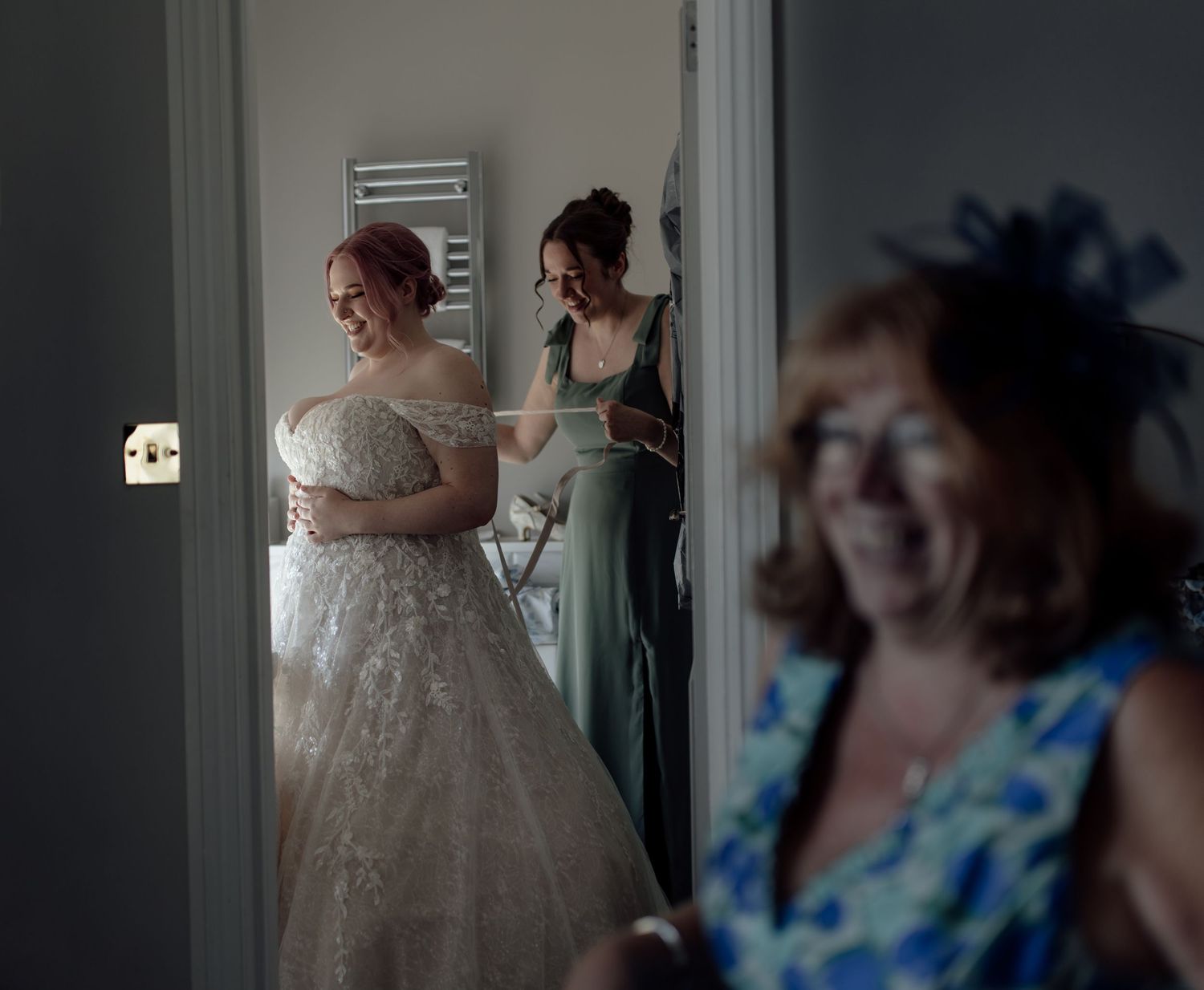 A bride in a white lace wedding dress stands in a doorway while someone in a sage green dress helps with preparations.