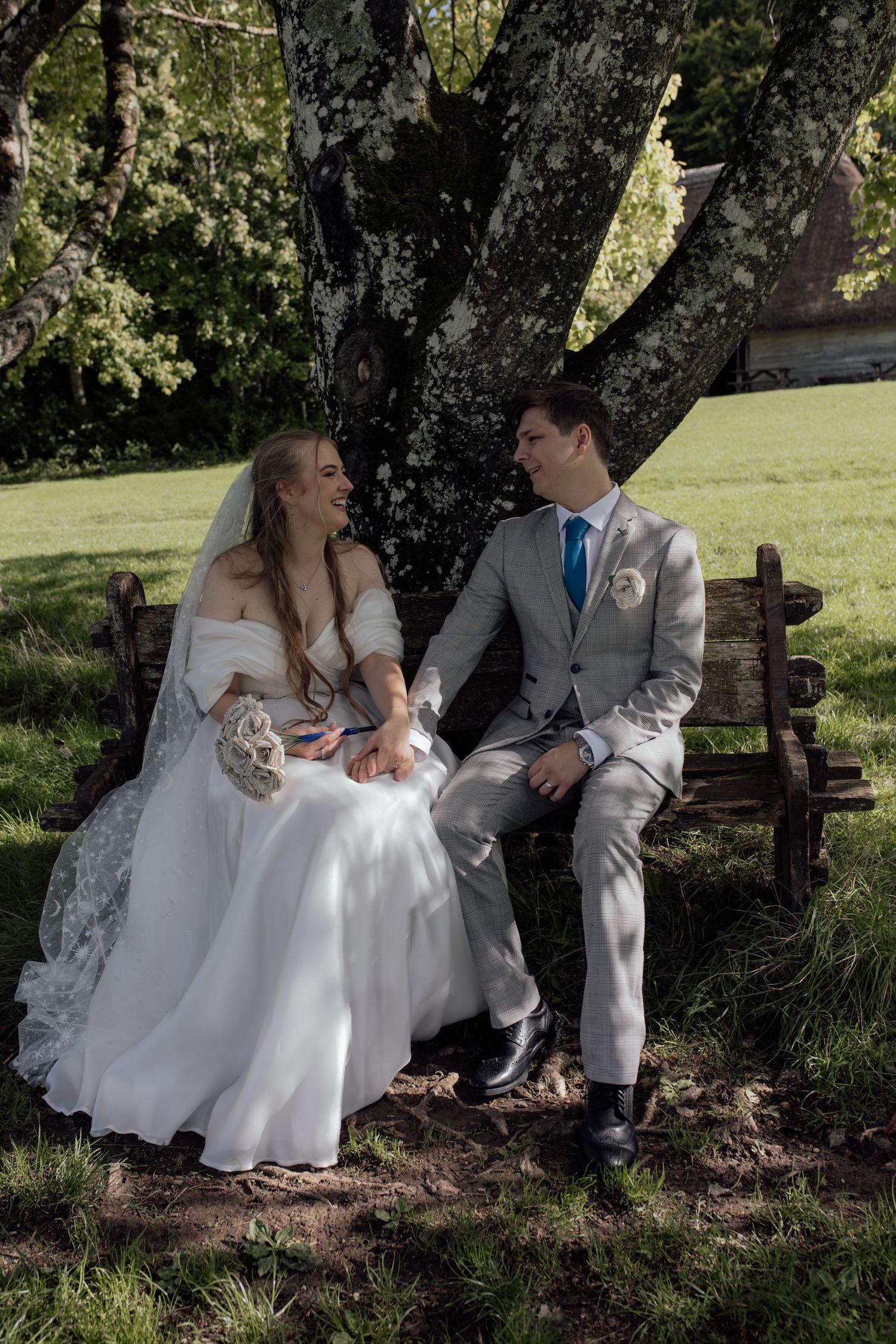 Bride and groom sit on bench holding hands, looking at each other.