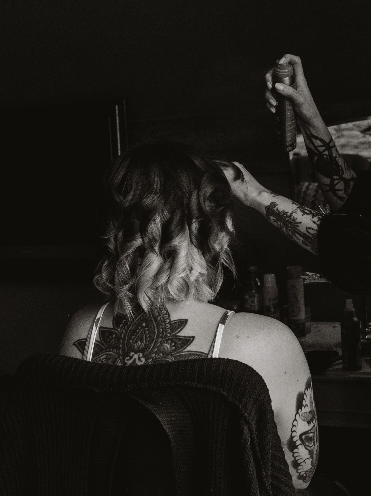 A black and white photo shows the back of someone's curled hair and shoulder with visible tattoos in a dark setting.