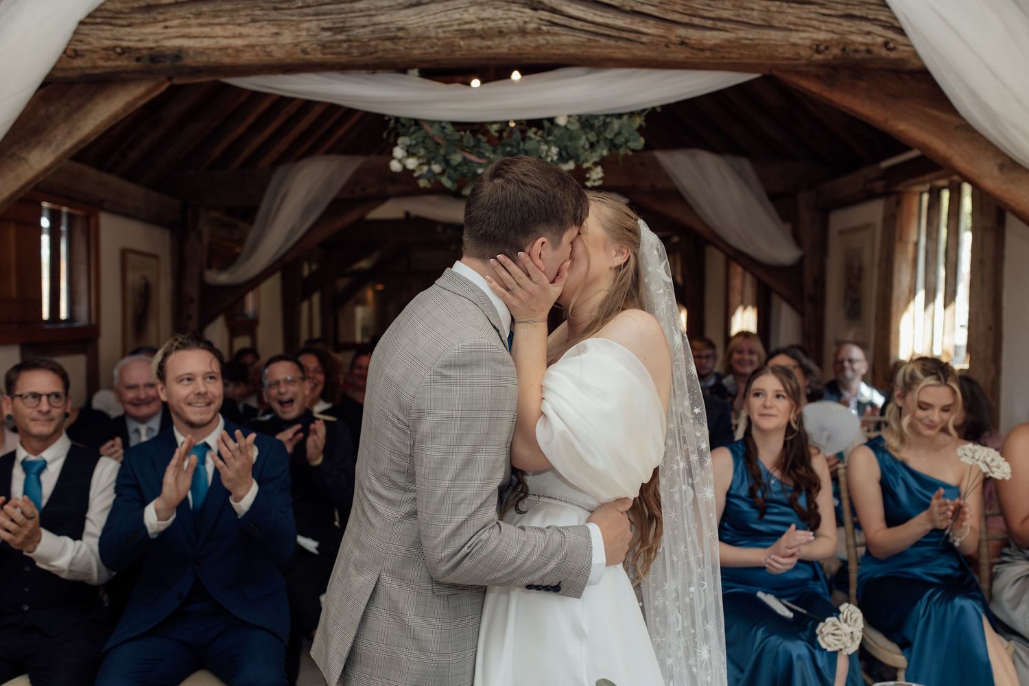 Newlyweds share their first kiss surrounded by guests at a rustic barn wedding reception.