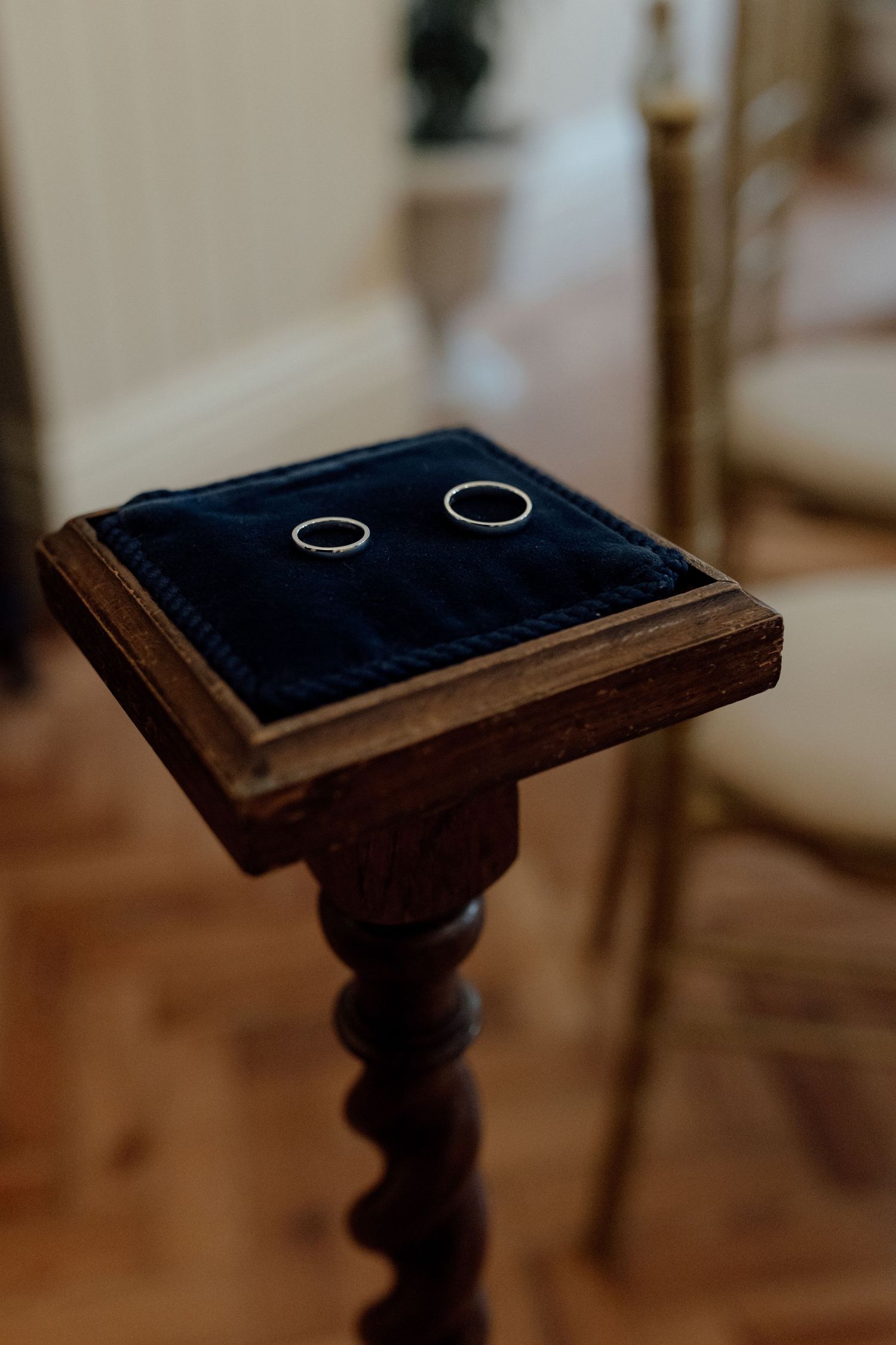 Two silver wedding rings rest on a dark velvet cushion atop an ornate wooden pedestal in warm lighting.