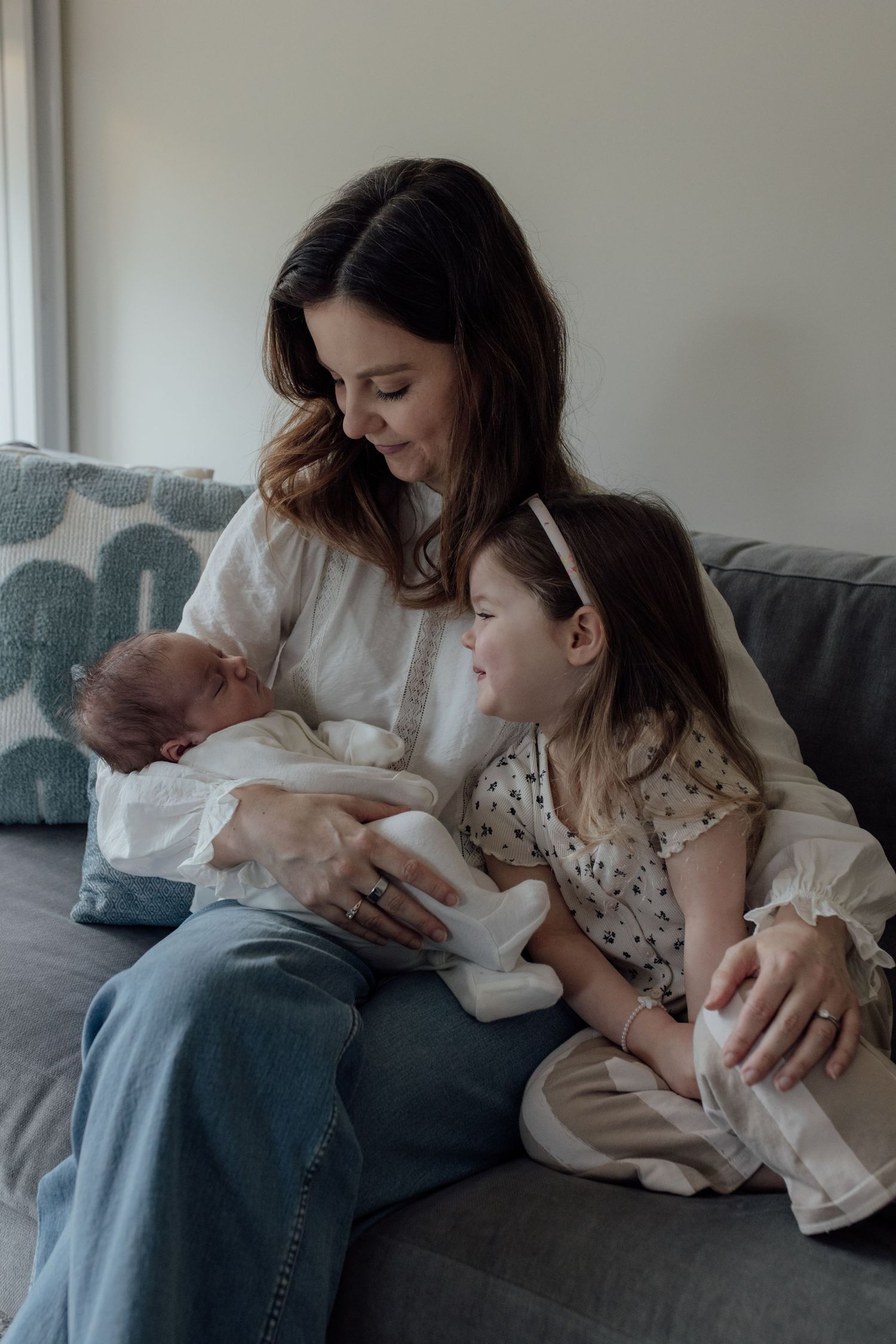 Family sits together on grey couch in intimate series of lifestyle photos showing tender moments at home.