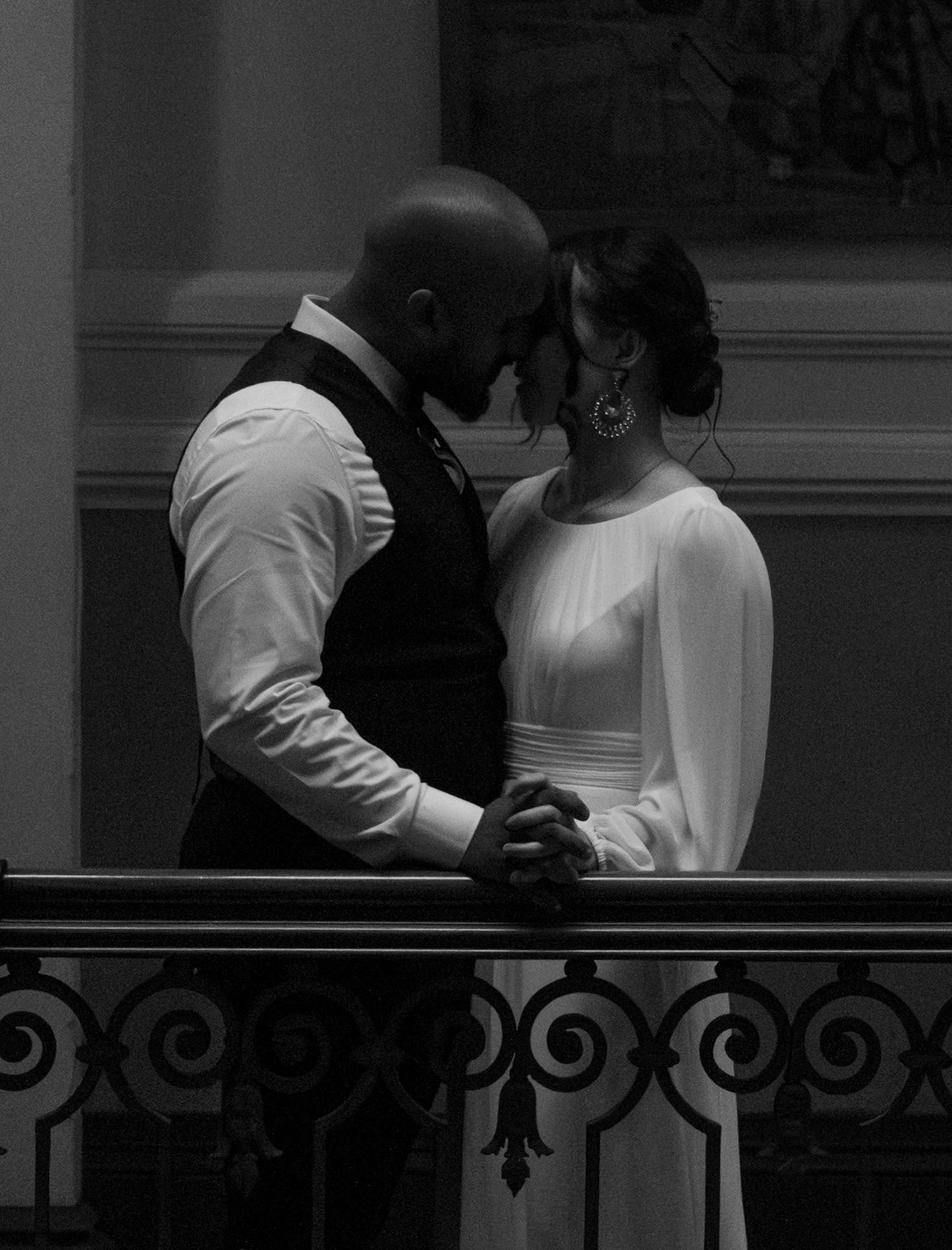 A couple in white clothing sits together on a bench behind decorative metal railings.