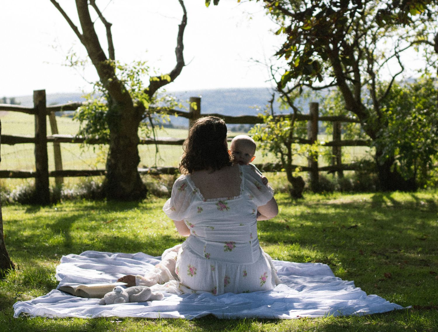A person relaxes on a white blanket under a shady tree on a sunny summer day in a grassy field.