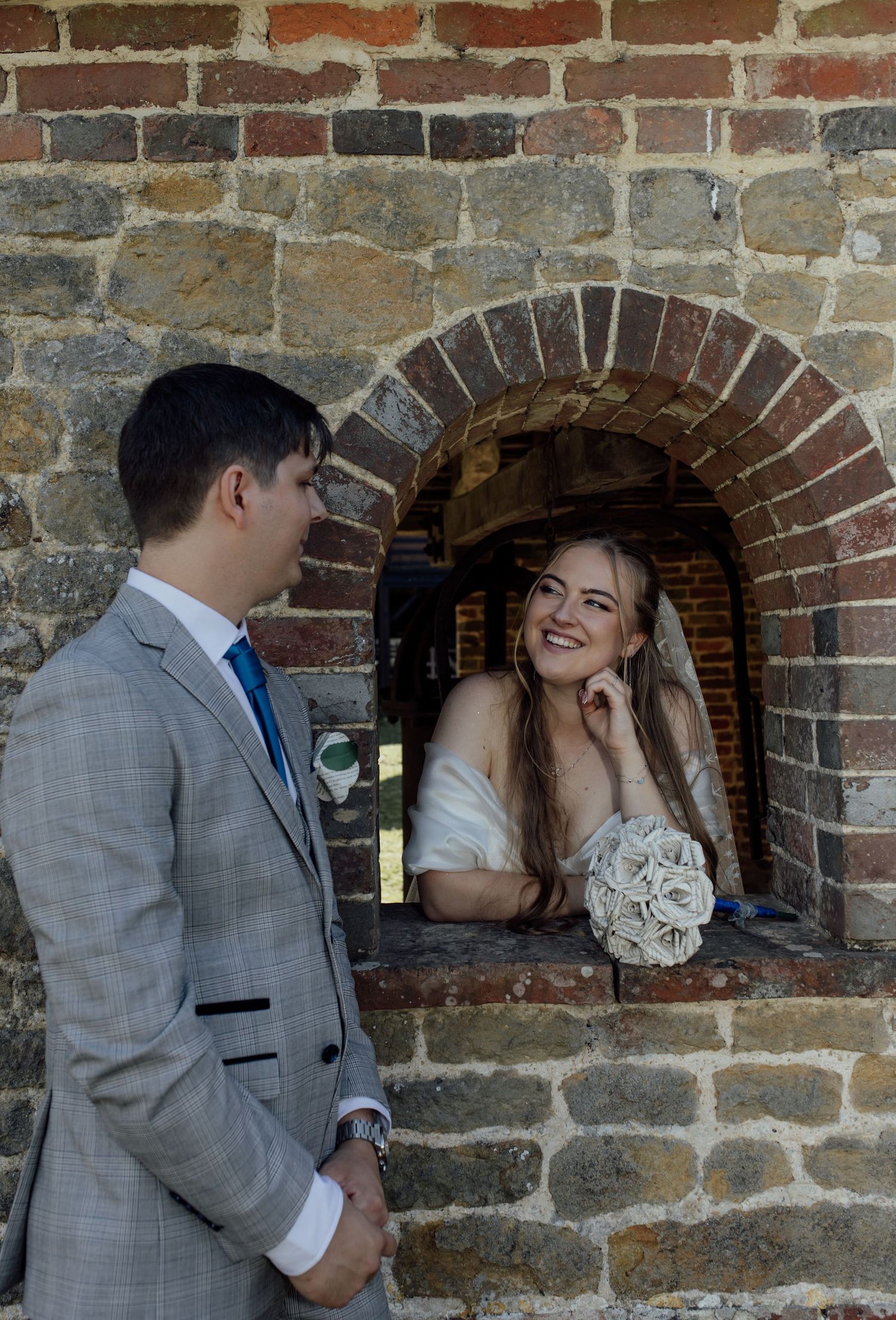 A couple in wedding attire shares a romantic moment against a rustic stone wall with an arched window.