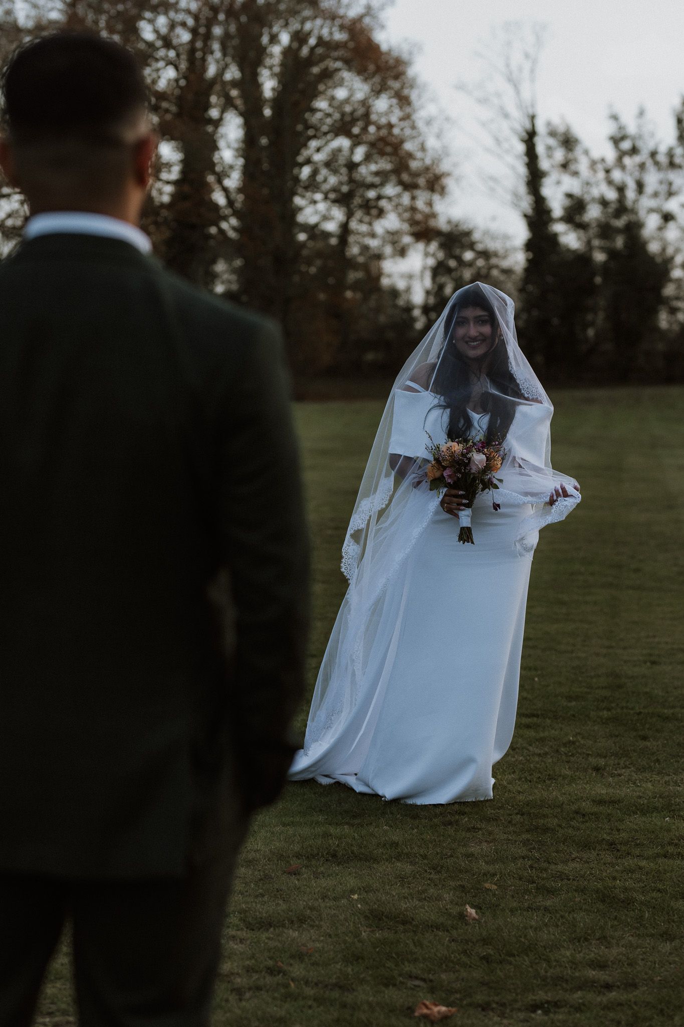 A bride in a flowing white dress and veil holds a bouquet while standing on a grassy lawn.