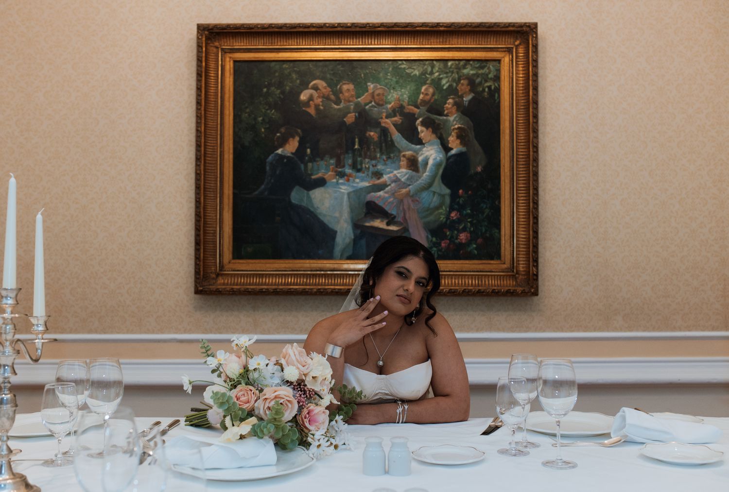A formal dining setting with white plates, candles, floral arrangements and a framed painting on the wall.