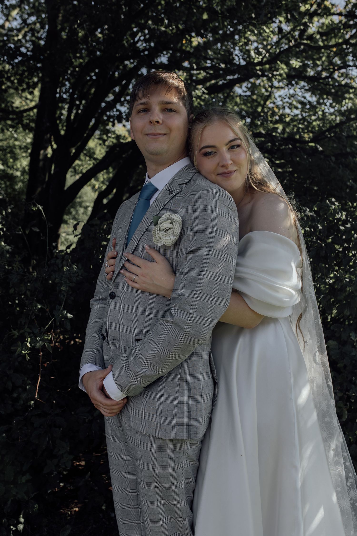 A couple in wedding attire embraces outdoors with trees in the background during their special day.
