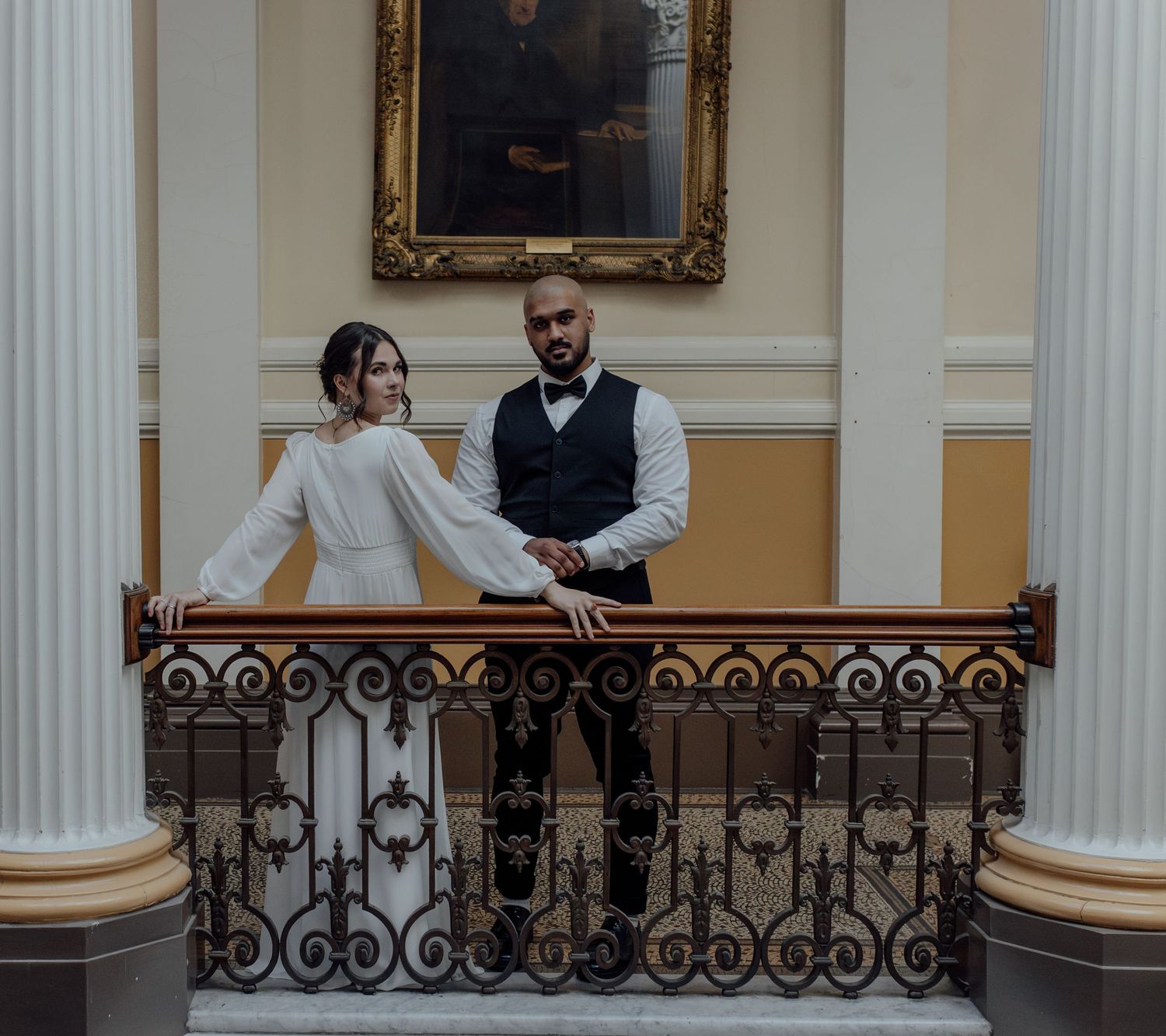 Couple shares a tender moment on an elegant staircase with ornate iron railings and white columns in a historic building.