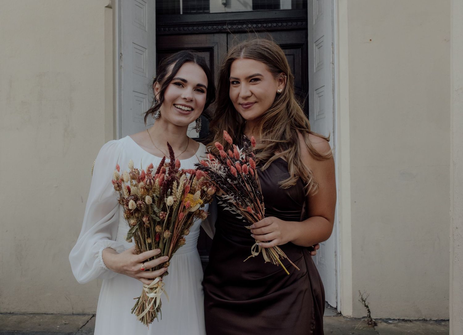 Two people stand together outdoors holding colorful autumn bouquets and smiling warmly.