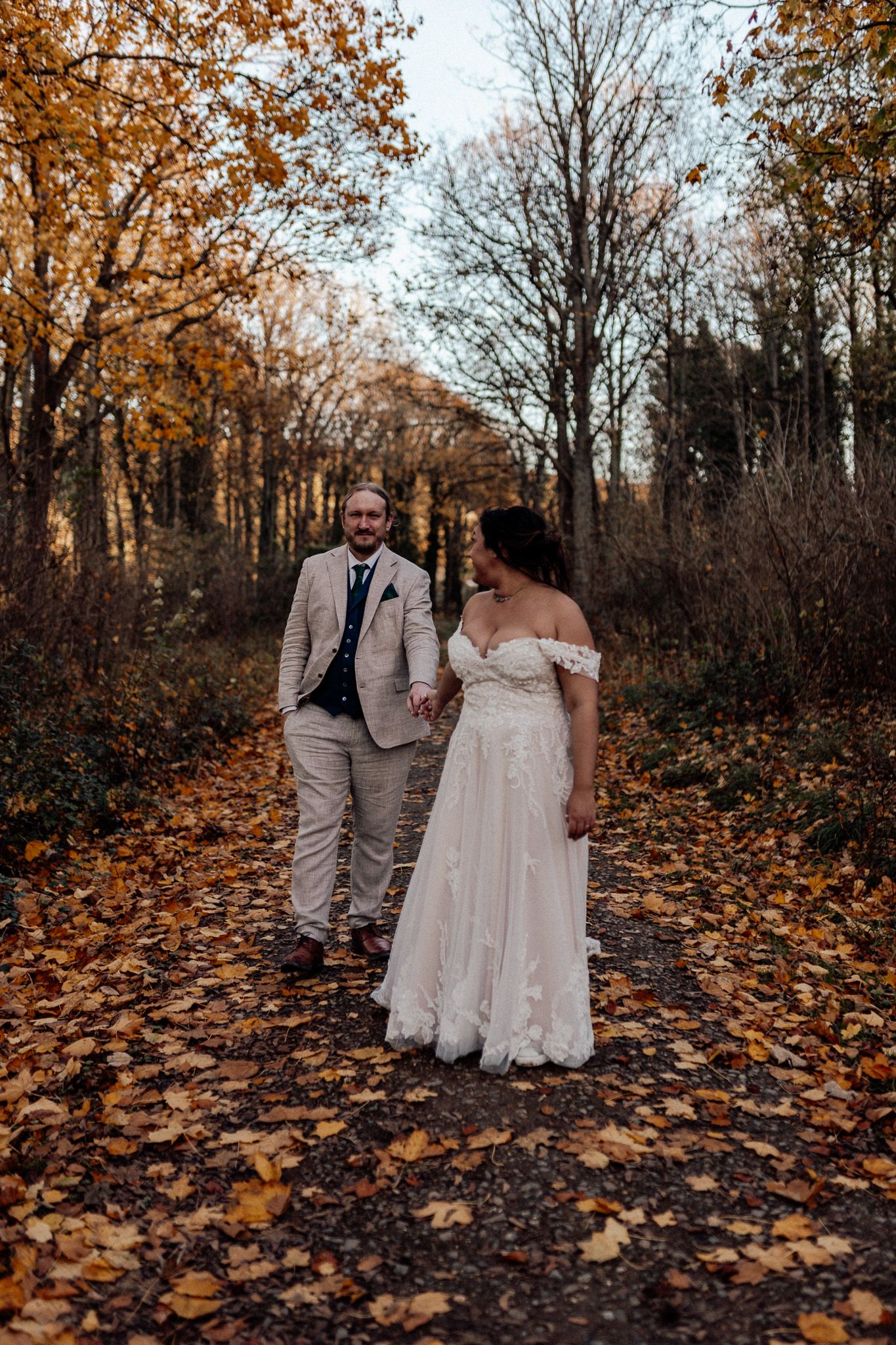 A couple in wedding attire walks through a forest path covered in golden autumn leaves during a fall photo session.