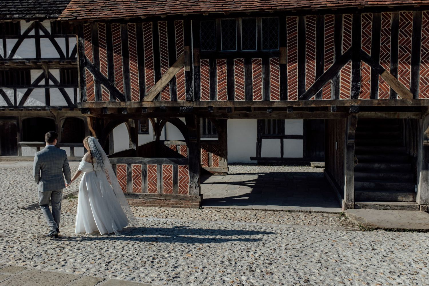 A couple in wedding attire walks through a rustic Tudor-style courtyard with timber-framed buildings.
