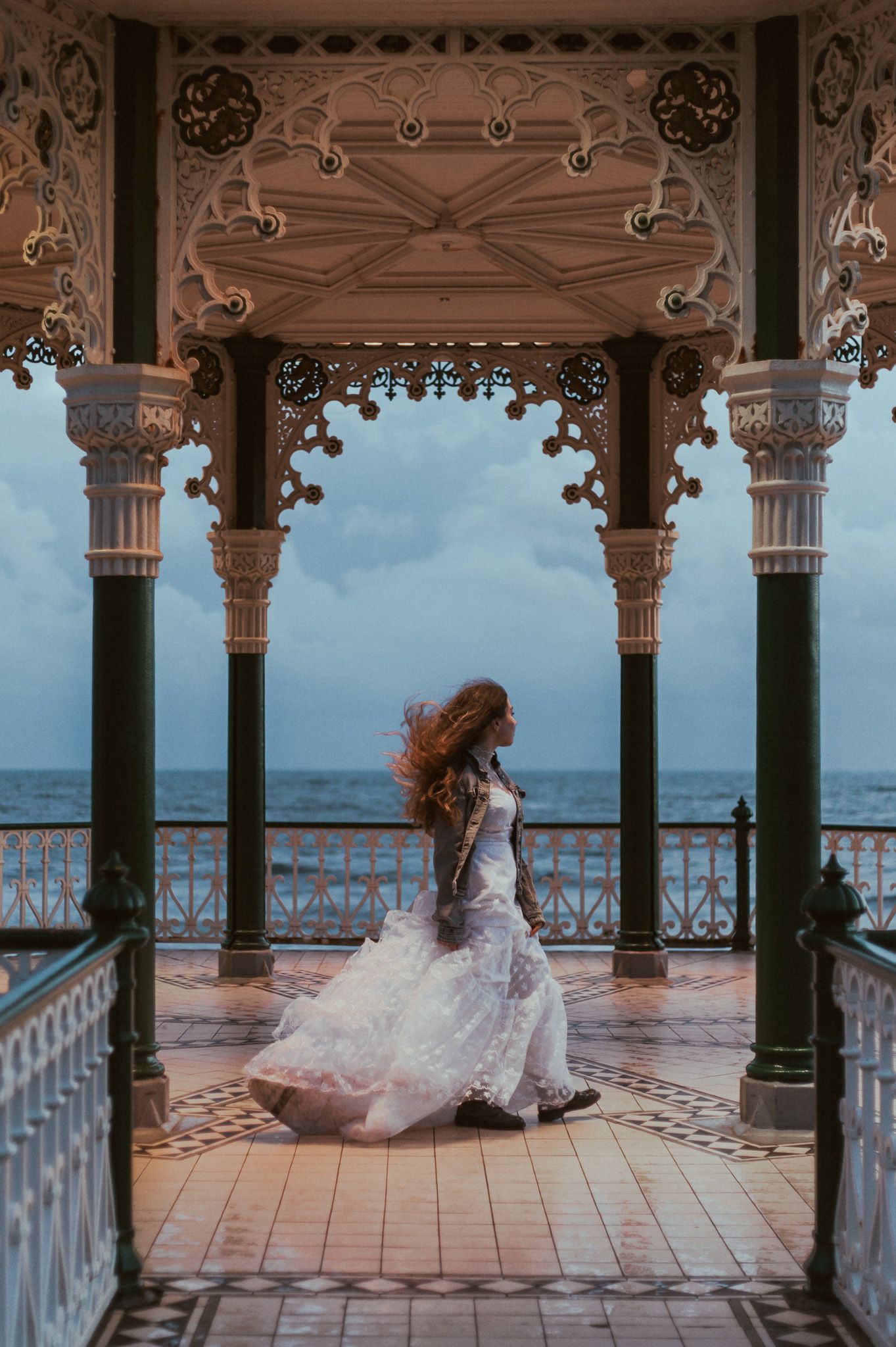 Figure in flowing white wedding dress twirls on an ornate Victorian-style balcony overlooking the ocean at dusk.