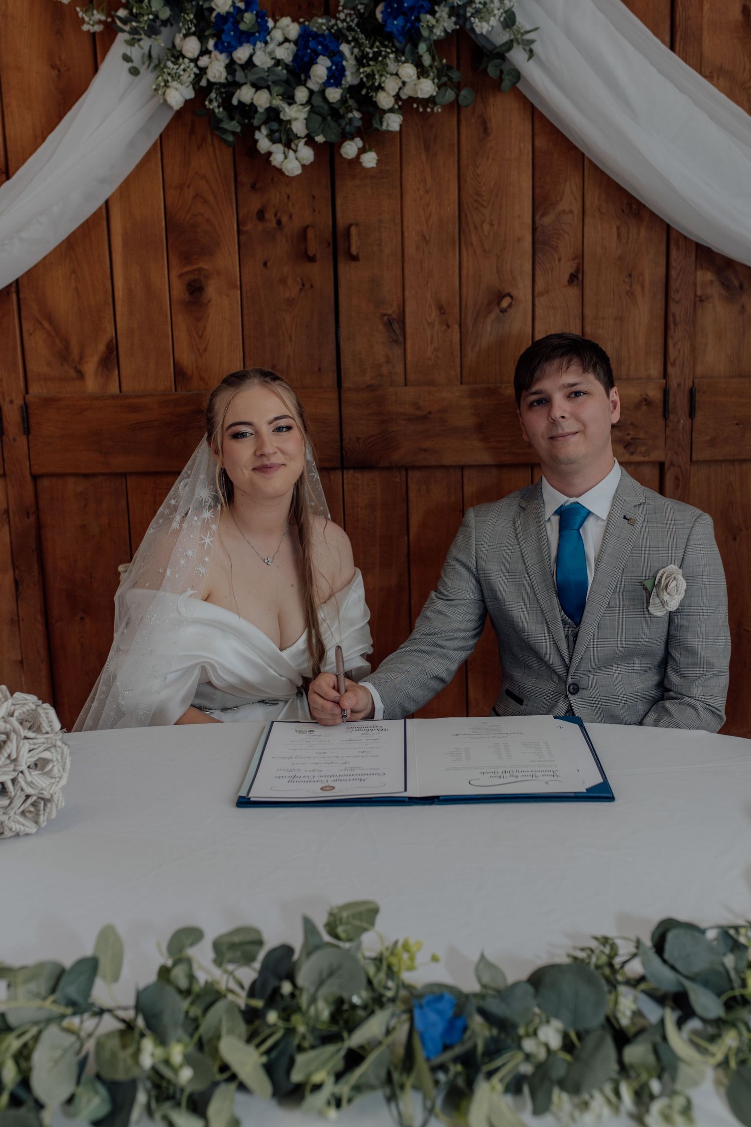 Couple signing marriage documents at rustic wooden table decorated with white flowers and eucalyptus.