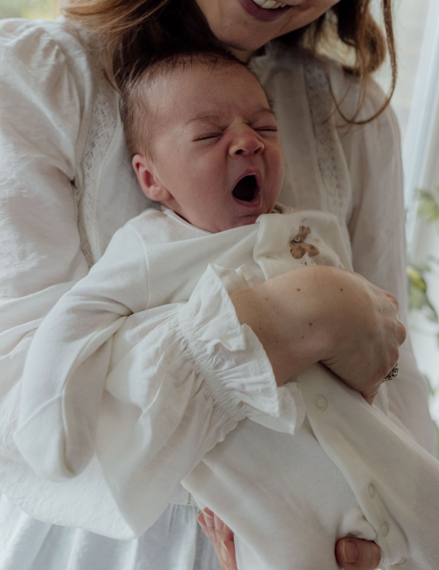 A baby in white clothing yawns while lying on white bedding.