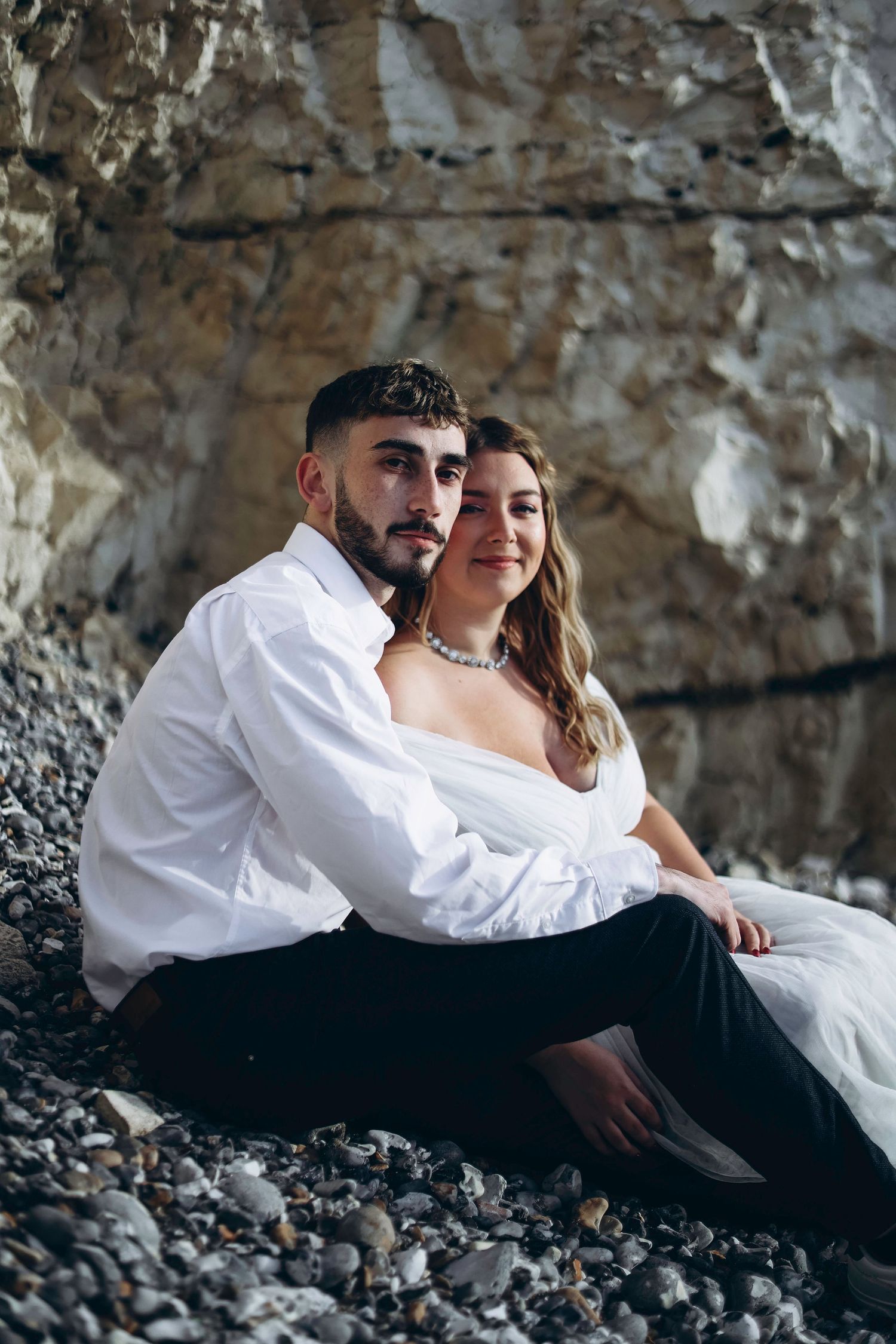 Couple in wedding attire sits together on rocky beach against dramatic cliff backdrop during romantic photoshoot.