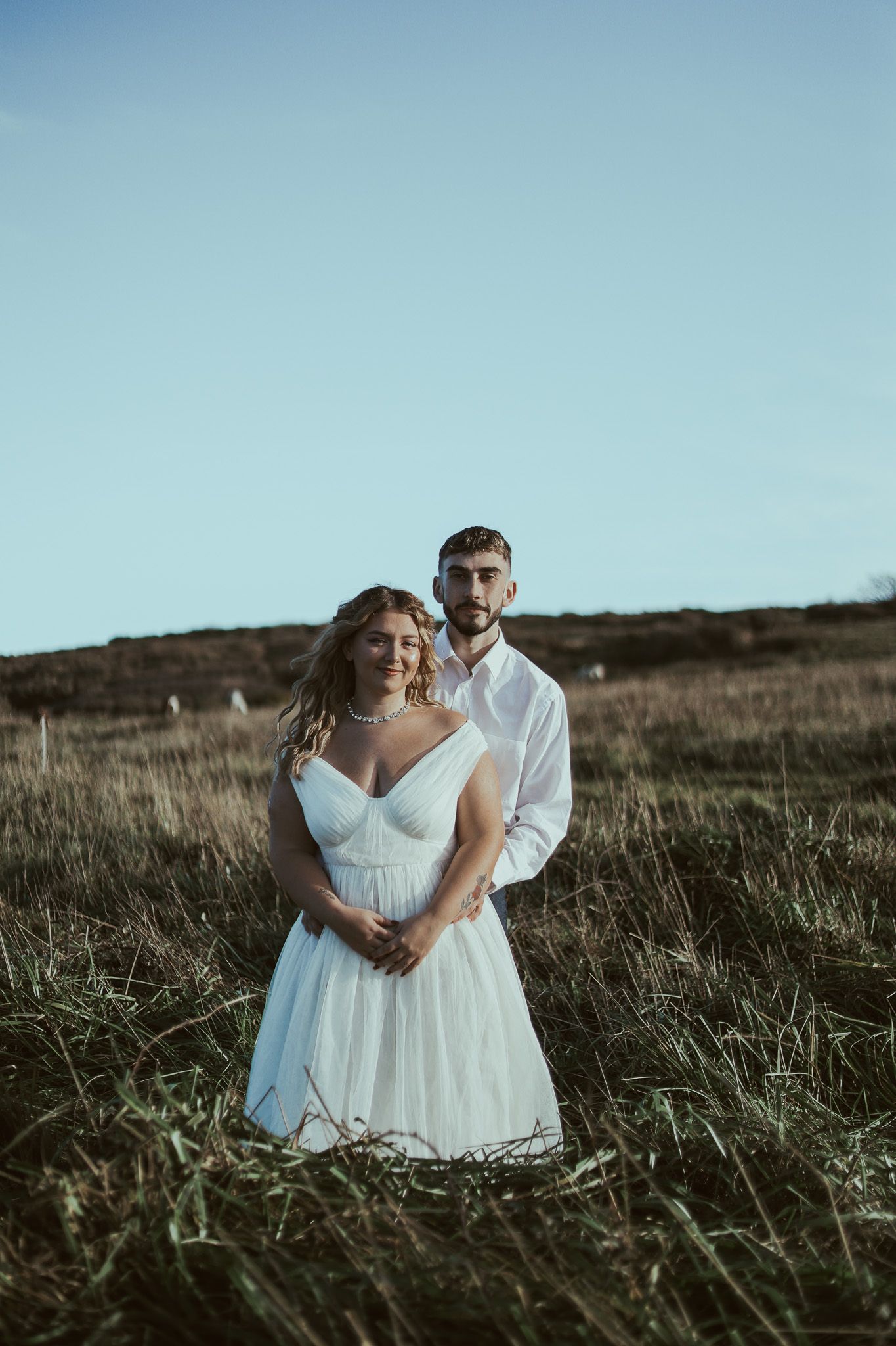 A couple in wedding attire embrace while sitting together in a sun-dappled grassy field at dusk.