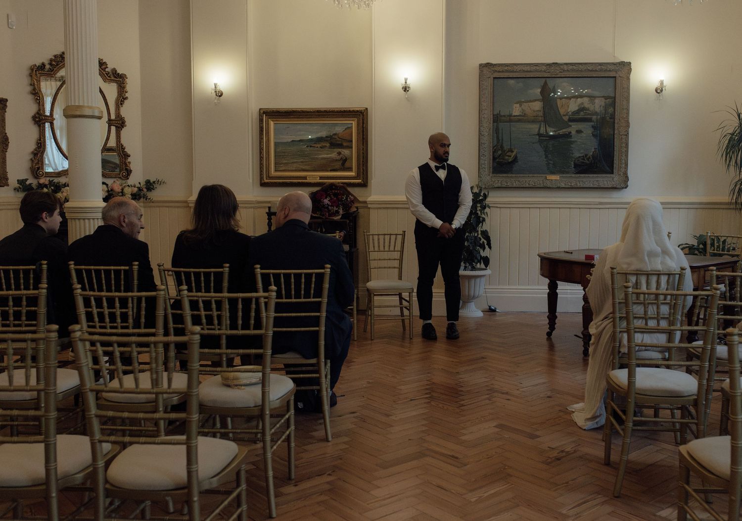 An elegant room with parquet flooring and gold chairs arranged for a ceremony, with wall sconces and framed artwork.