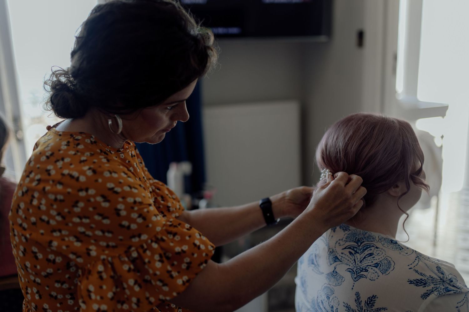 A stylist in an orange floral print blouse works on styling a client's hair near a window.