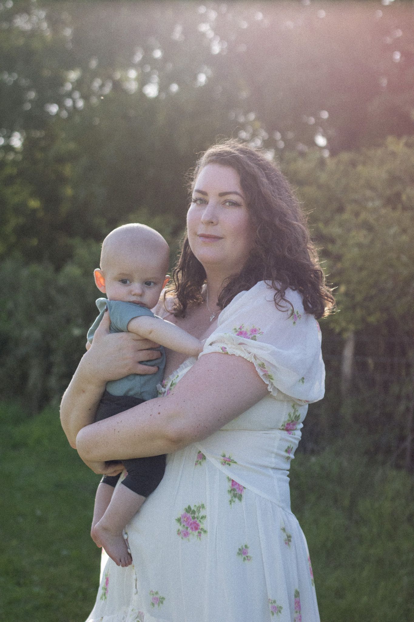 A woman in a white floral dress holds a baby outdoors at sunset with trees in the background.