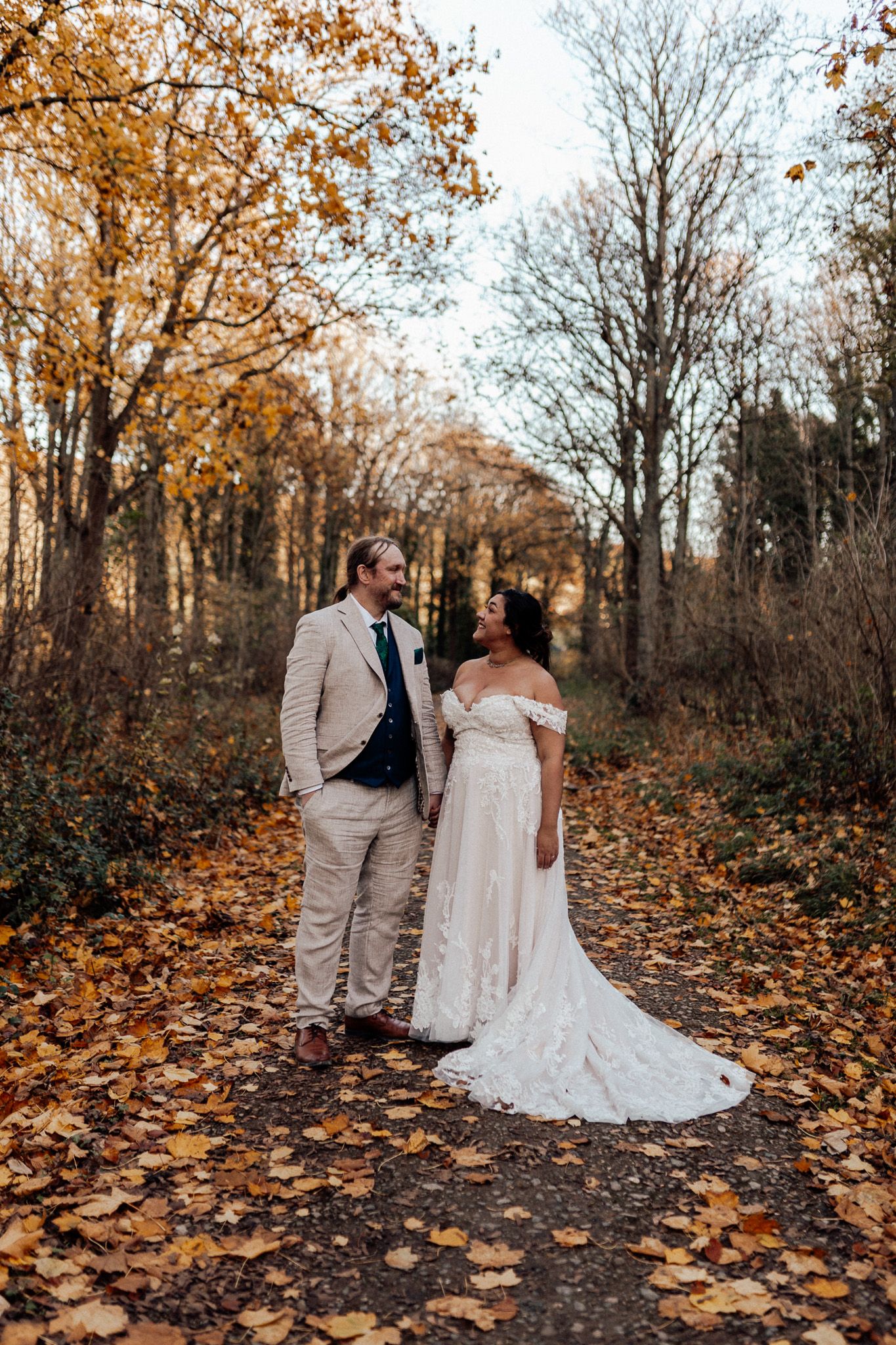 A couple in wedding attire shares romantic moments on a path covered in fallen autumn leaves during a fall photo session.