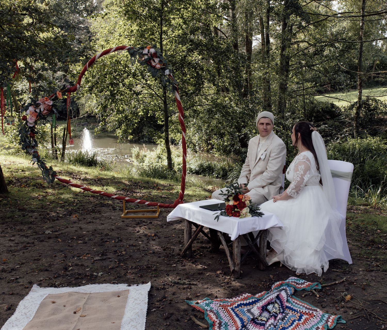 A wedding couple enjoys a rustic outdoor swing decorated with red ribbons and floral arrangements in a wooded setting.
