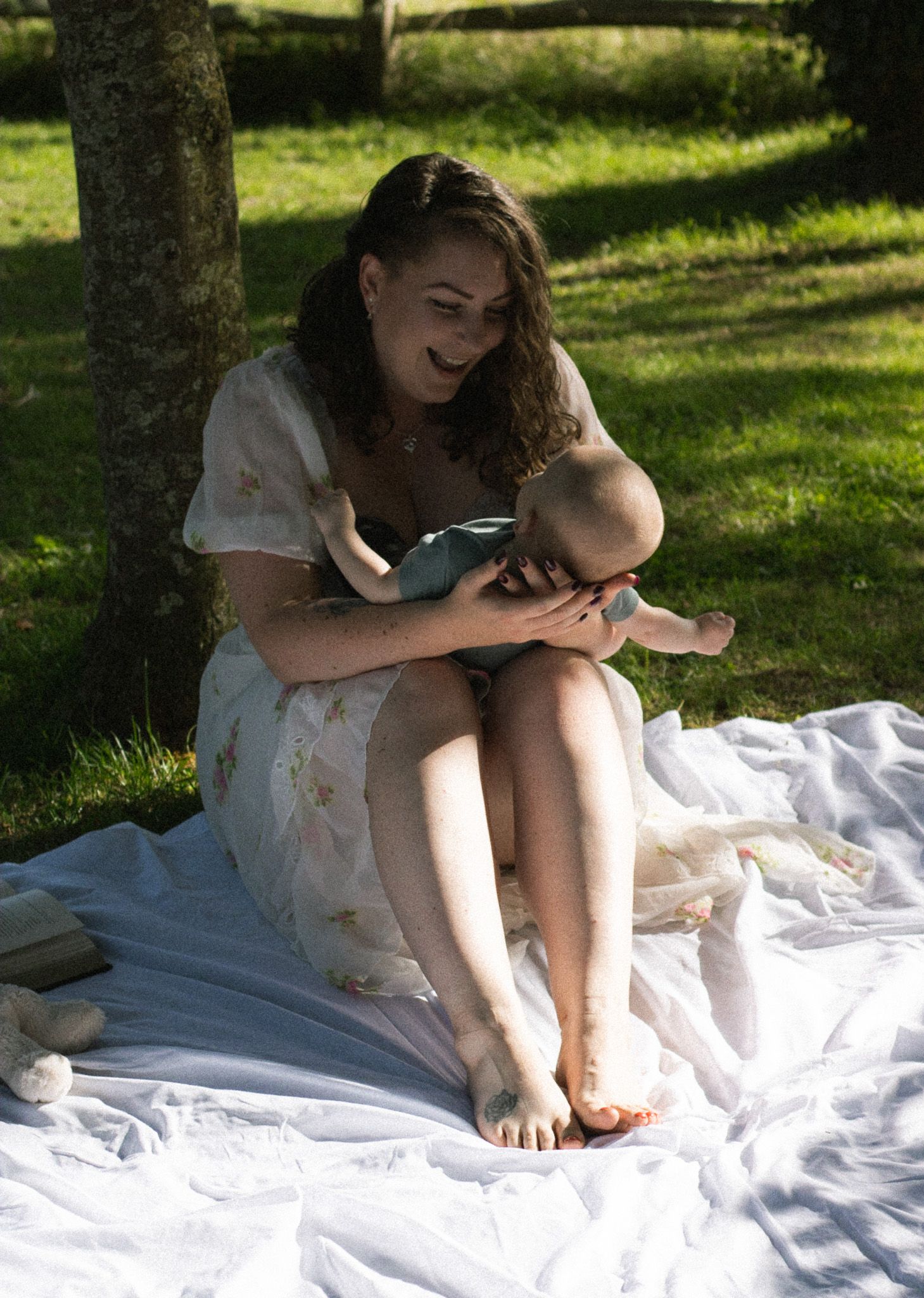 A tender moment captured outdoors on a white blanket under a tree on a sunny summer day.